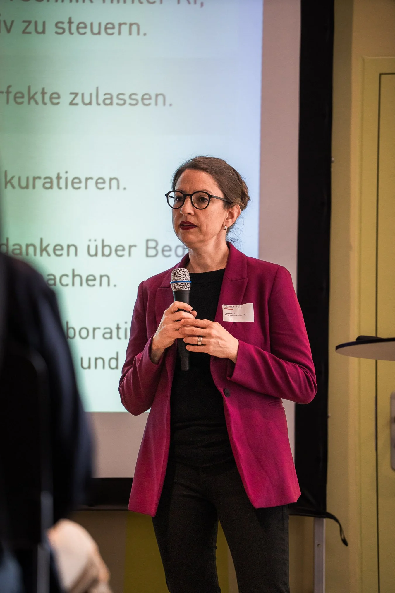 Frau mit Brille und roten Blazer hält Mikrofon bei Präsentation, im Hintergrund ein Bildschirm mit deutschem Text.