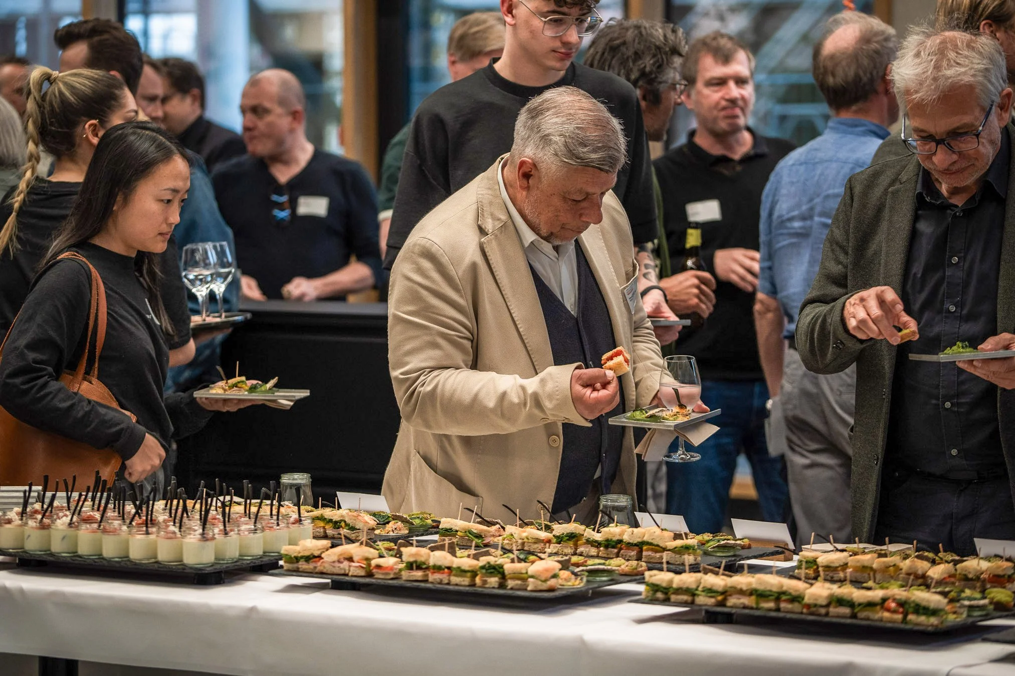 Menschen bei einem Empfang bedienen sich an einer großen Buffettafel mit Vorspeisen und Häppchen in einem modernen Raum.