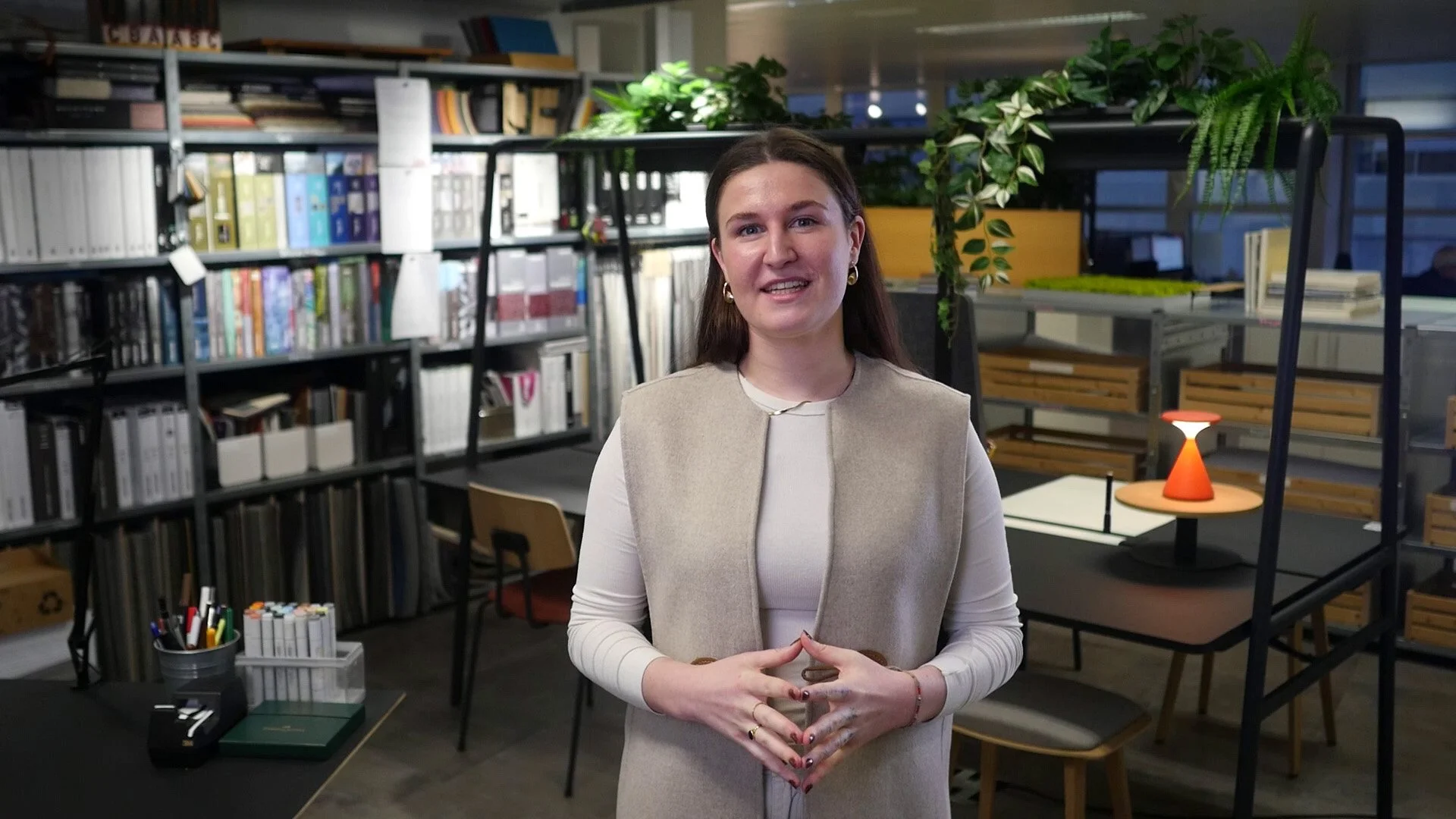 Eine Frau steht in einem Raum mit Büchern und Pflanzen, vor einem Tisch mit Schreibtubehör, während sie direkt in die Kamera lächelt.