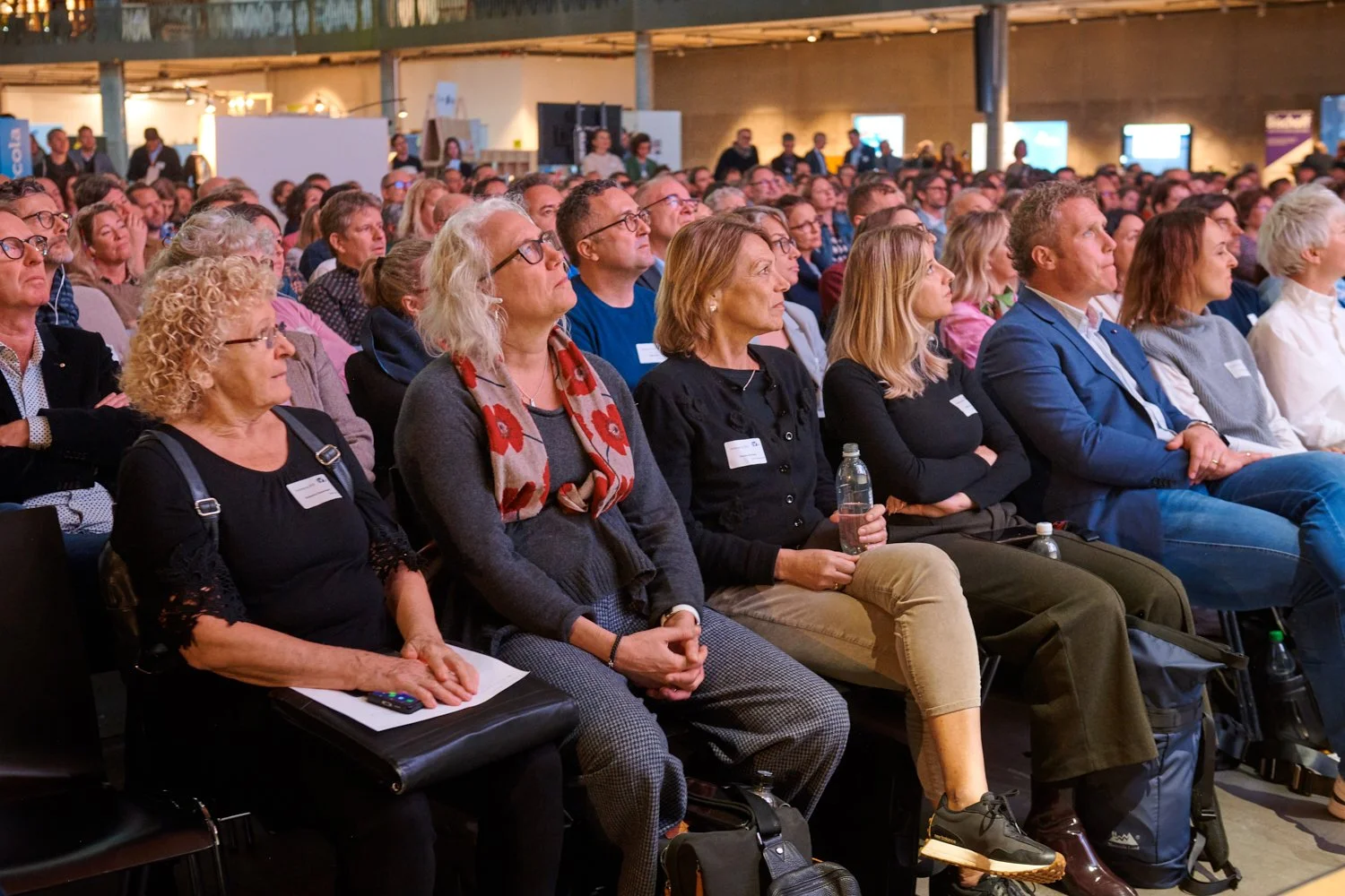 Eine Gruppe von Menschen sitzt in einem großen Raum bei einer Konferenz oder einem Vortrag, die meisten schauen aufmerksam nach vorne.