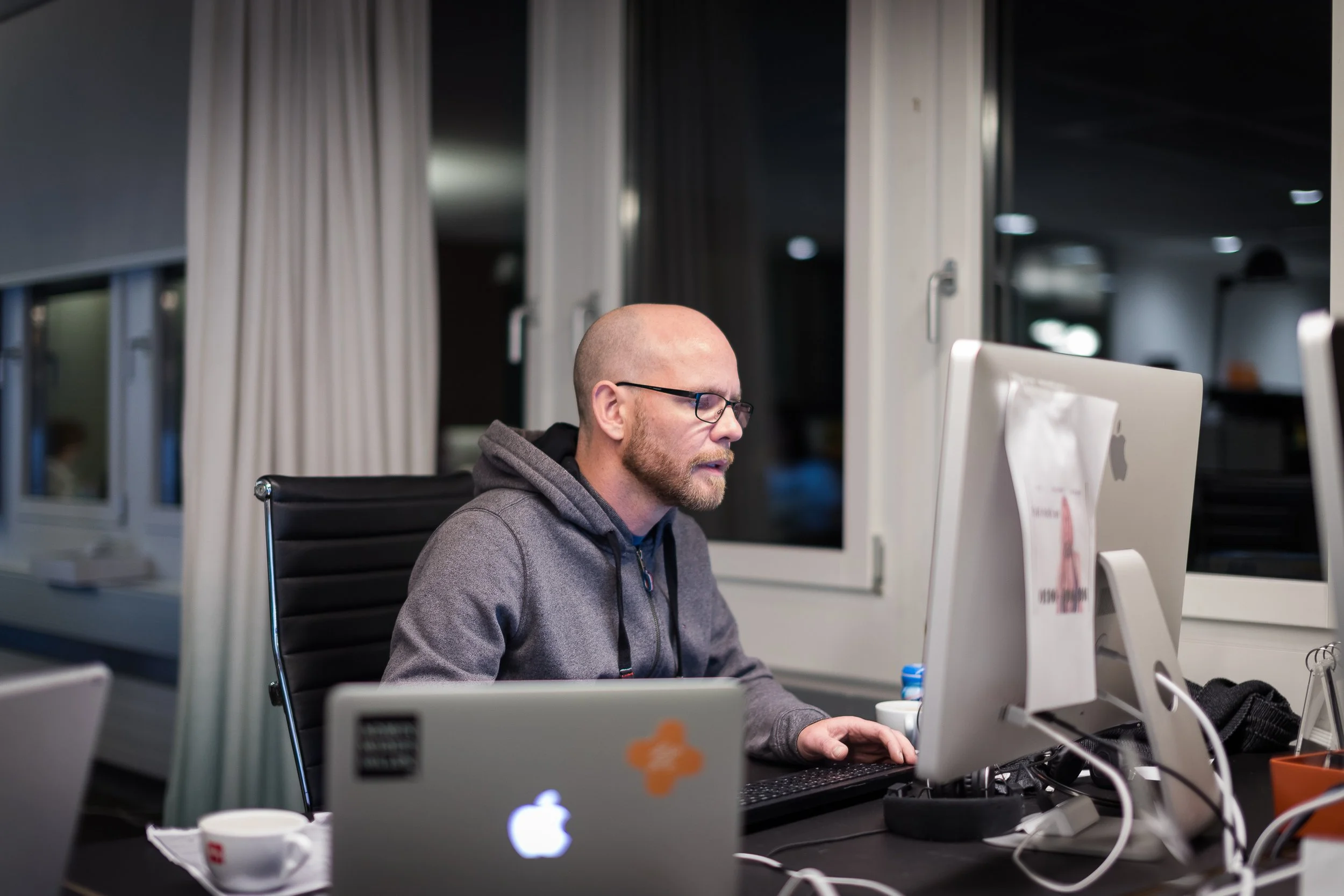 Mann mit Brille arbeitet an einem Computer in einem Büro, umgeben von weiteren Computern und Tassen.