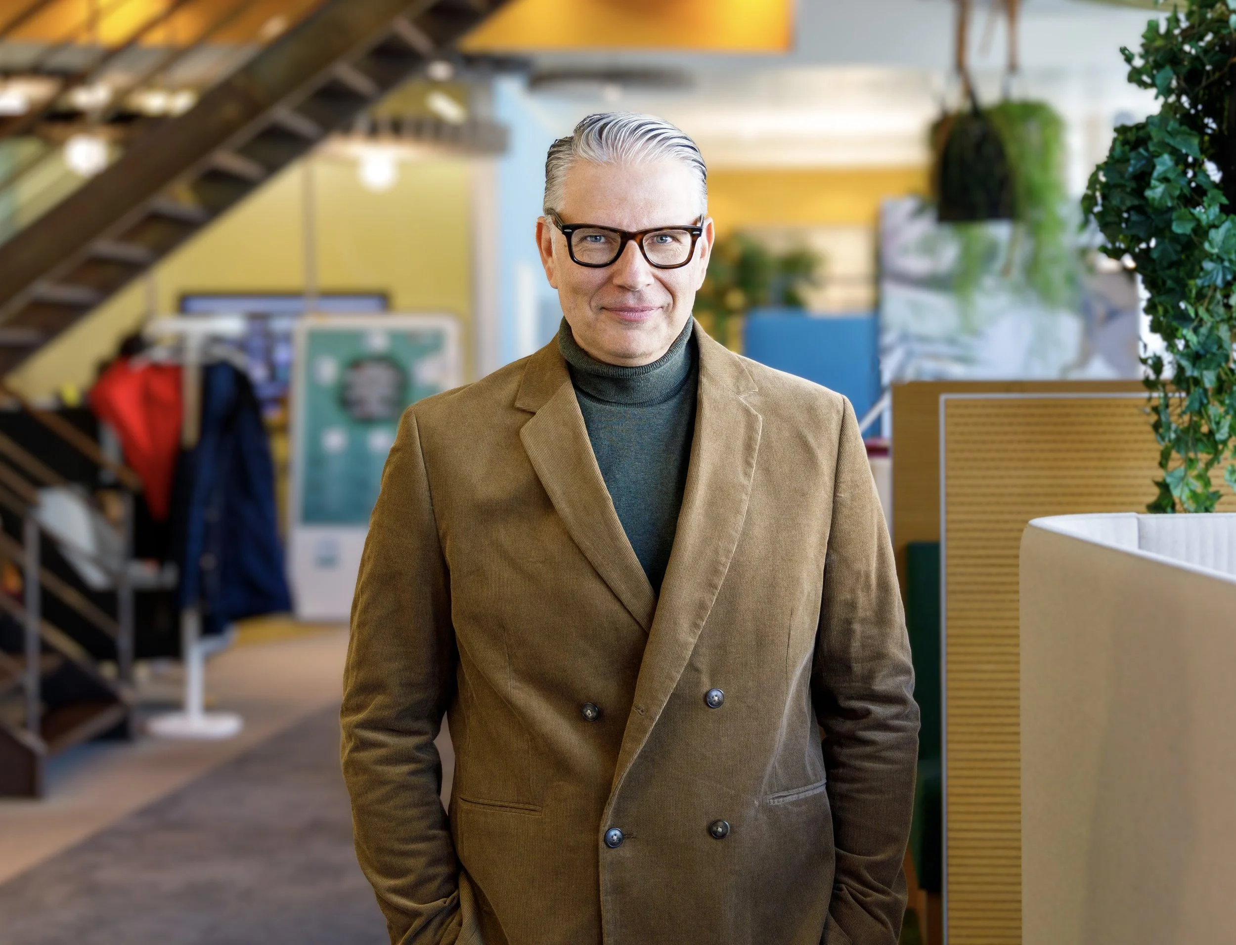 Ein Mann mit grauem, seitlich gescheiteltem Haar und schwarzen Brillen steht in einem modernen Büro, trägt einen braunen Blazer und einen schwarzen Rollkragenpullover, im Hintergrund sind Pflanzen und Büromöbel zu sehen.