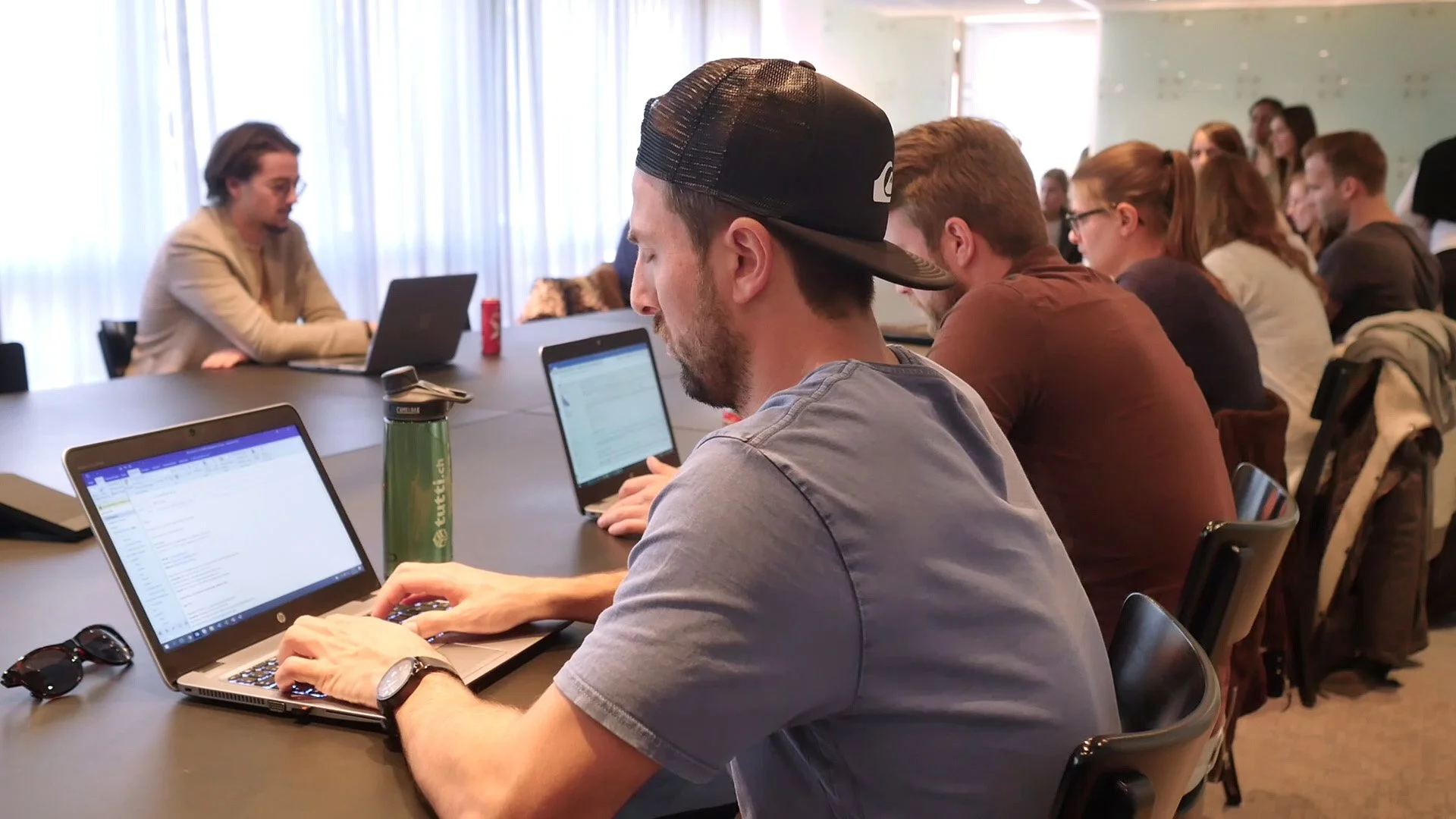 Menschen sitzen in einem modernen Konferenzraum mit großen Fenstern, die Tageslicht ins Zimmer lassen, und arbeiten an Laptops. Im Vordergrund eine Person in Grau mit schwarzer Kappe, im Hintergrund weitere Teilnehmer, die aufmerksam zuhören.