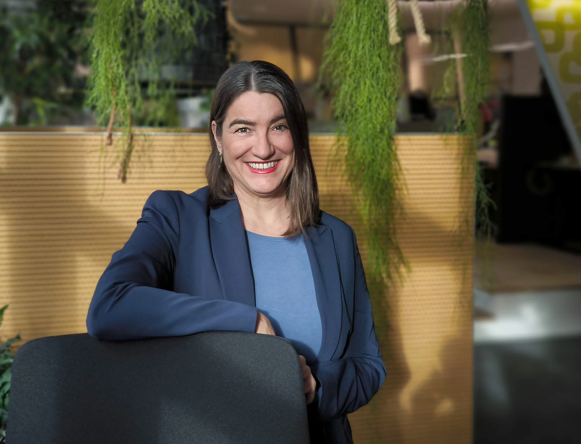 Lächelnde Frau in blauer Jacke steht an einem Bürostuhl vor grünem Hintergrund in einem modernen Raum.