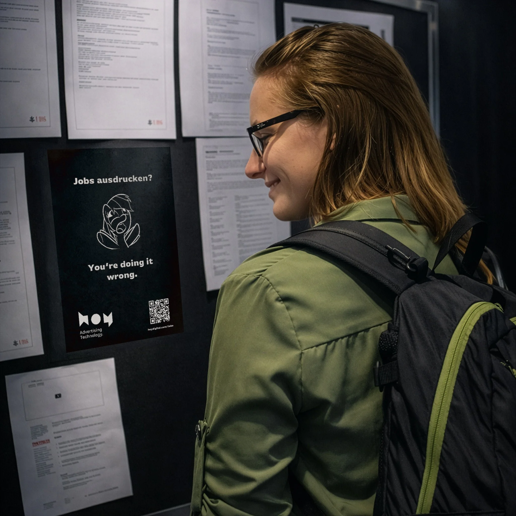 Eine junge Frau mit Brille, grünem Mantel und Rucksack liest aufmerksam eine Informationswand mit mehreren Dokumenten und Postern. Auf einem der Poster steht auf Englisch: "Jobs ausdrucken? You’re doing it wrong."