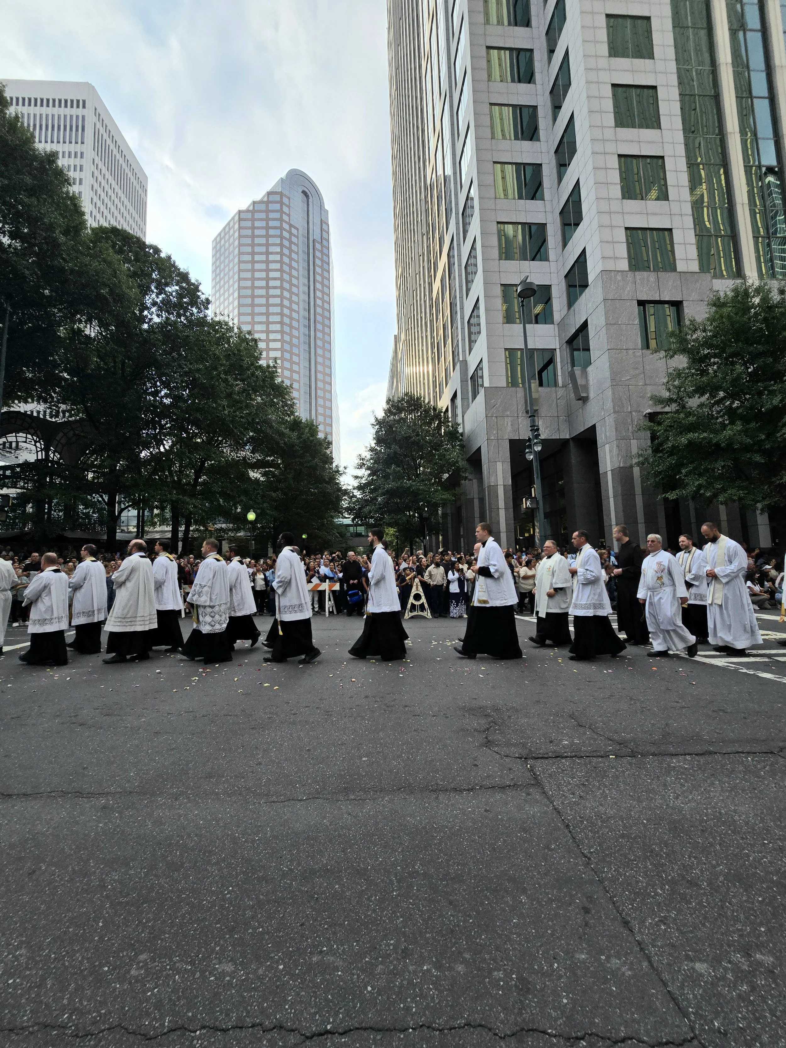 Clergy members in white and black robes walking in a procession on a city street with tall modern buildings and a crowd of spectators in the background.
