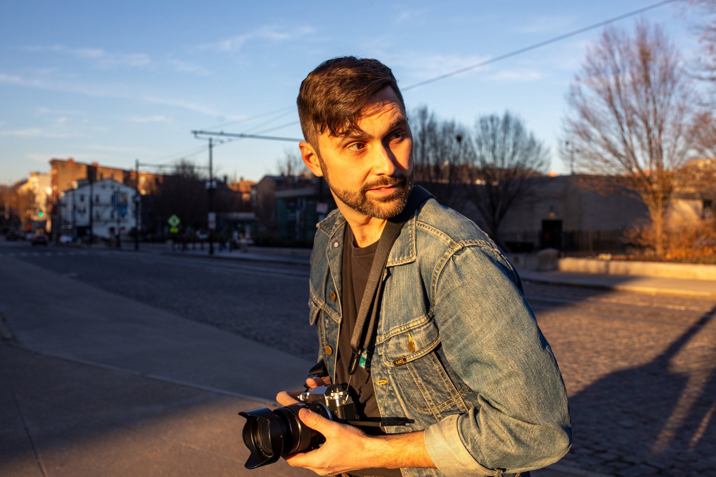 A man holding a camera outdoors during sunset, wearing a denim jacket with a black shirt underneath, and looking to his right.
