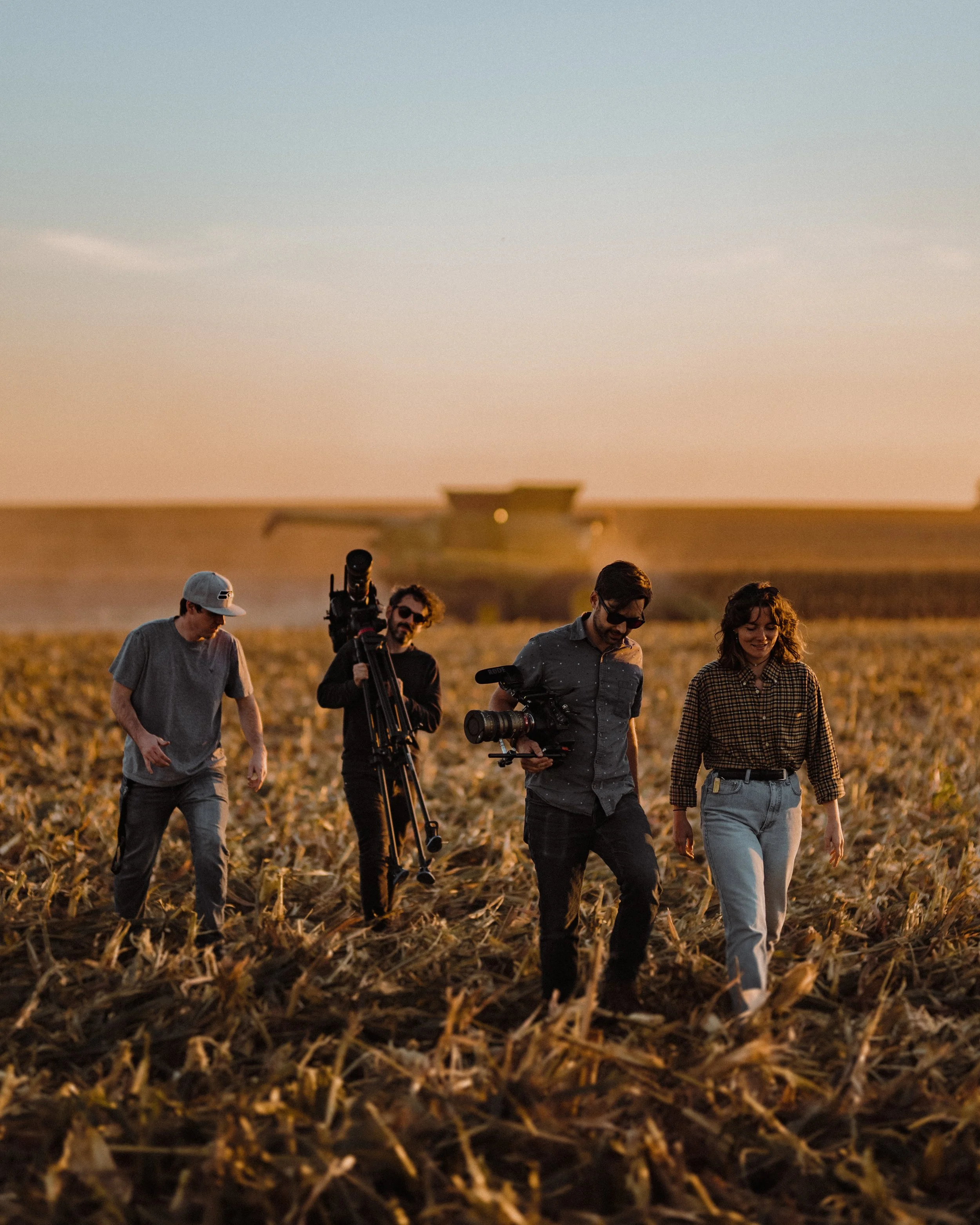 Four people walking through a field during sunset, with a combine harvester in the background and one person carrying a camera.