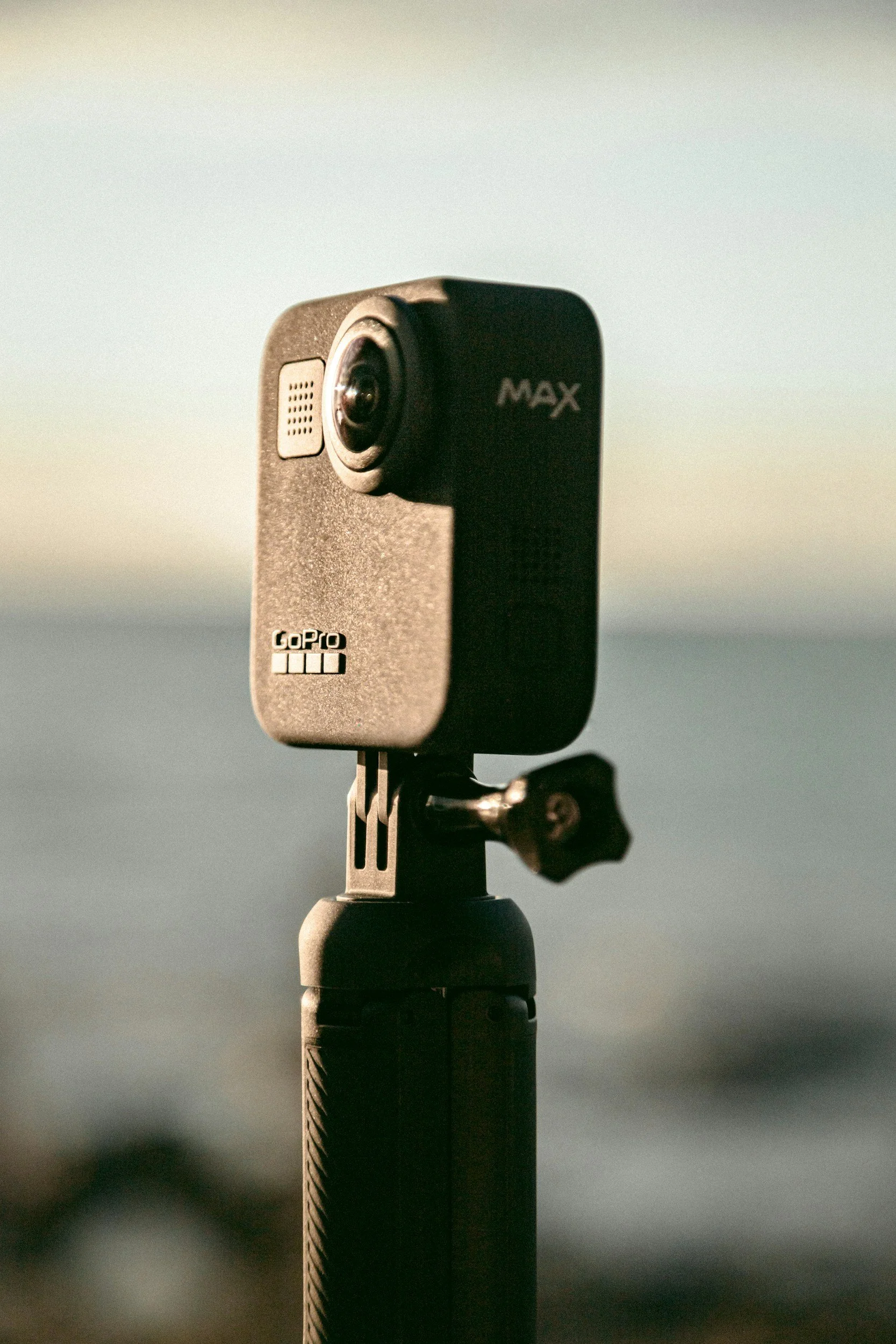 Close-up of a black GoPro MAX camera mounted on a monopod, against a blurred outdoor background.