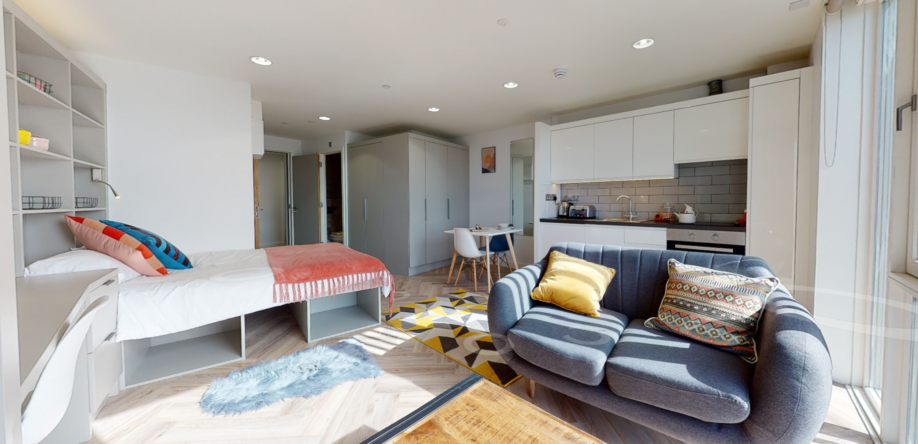 Studio apartment with a bed, sofa, kitchen area, and small dining table, decorated with colorful cushions and a geometric rug.