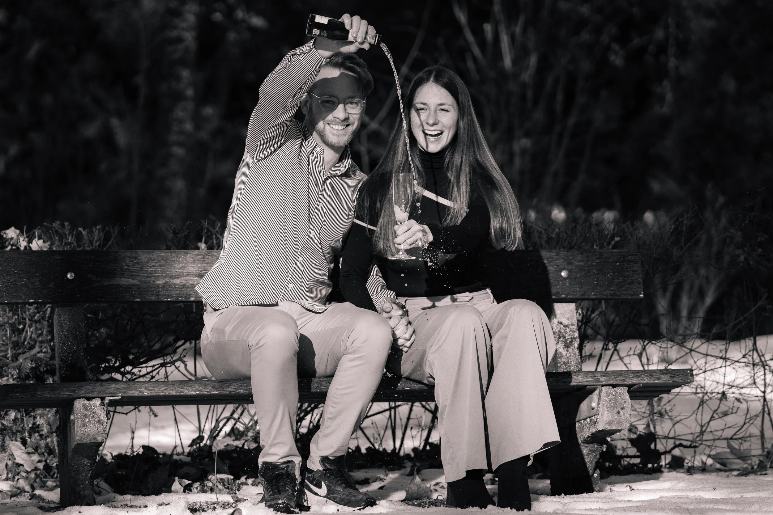 Ein lachendes Paar sitzt auf einer Parkbank im Schnee, trinkt Champagner, während sie eine Flasche öffnen, bei Nacht im Freien.