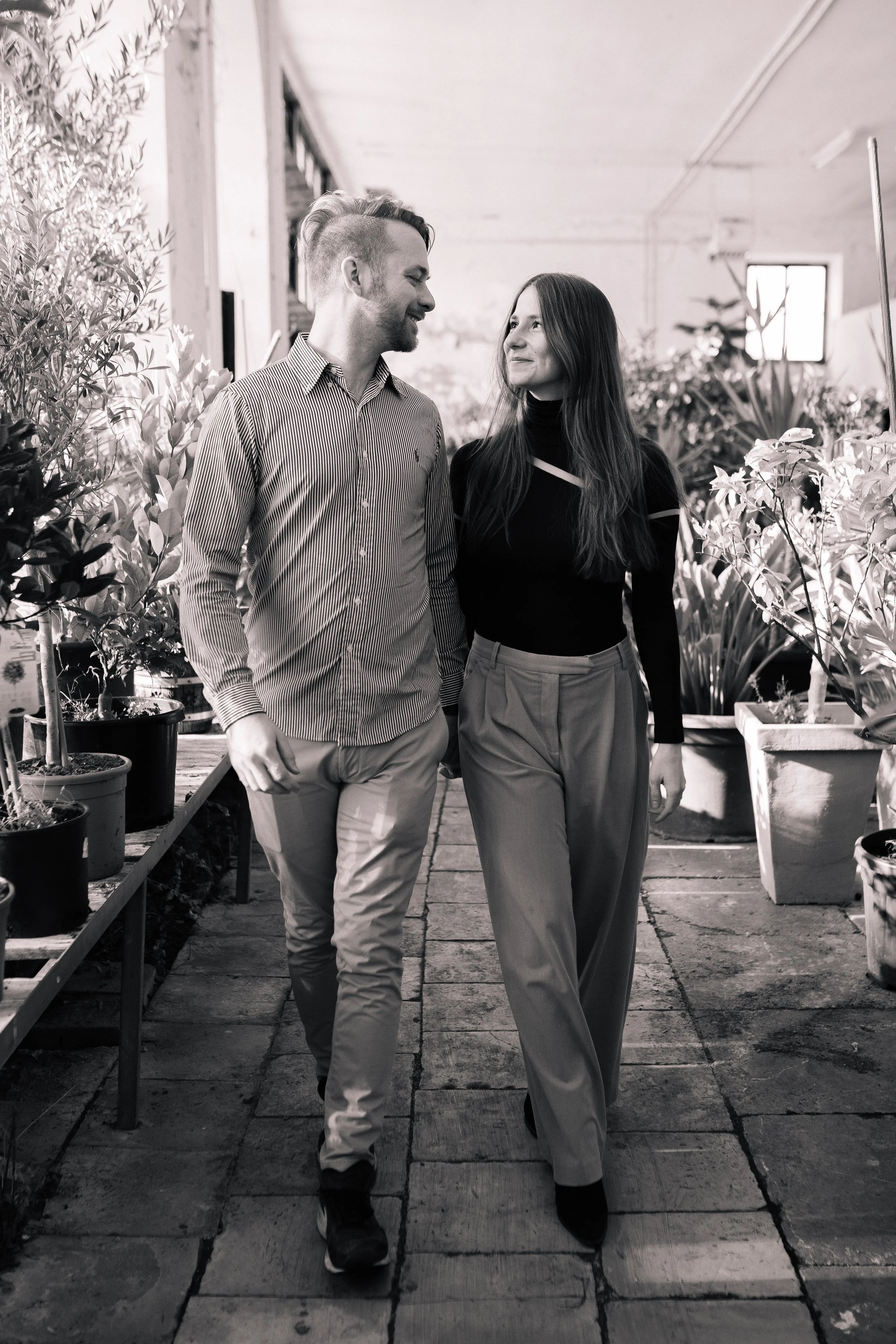 Ein Mann und eine Frau gehen Arm in Arm durch eine Gewächshaus mit Pflanzen, lächelnd und in einander vertieft.