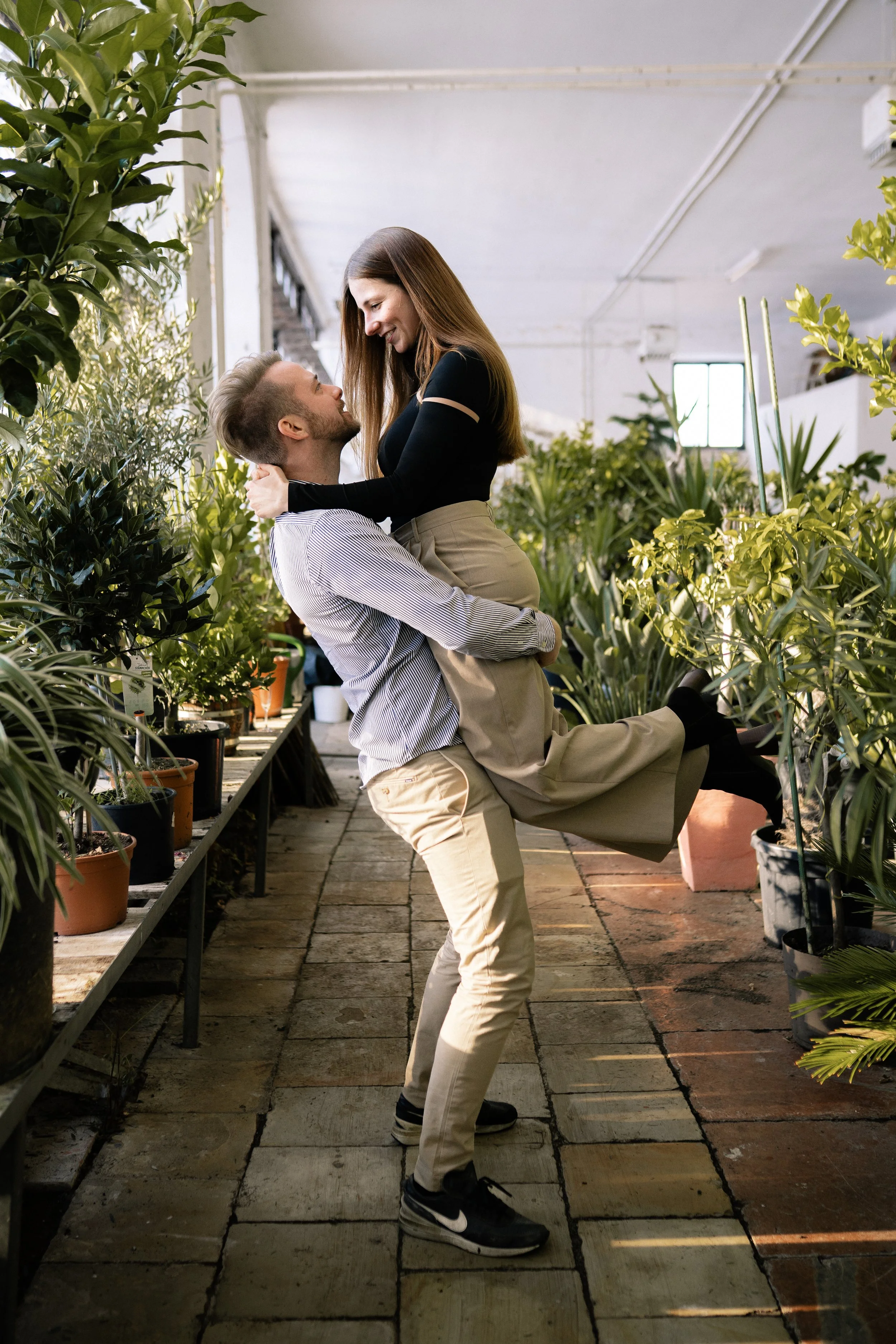 Ein lachendes Paar im Pflanzenladen, er hebt sie hoch zwischen Grünpflanzen und Töpfen.