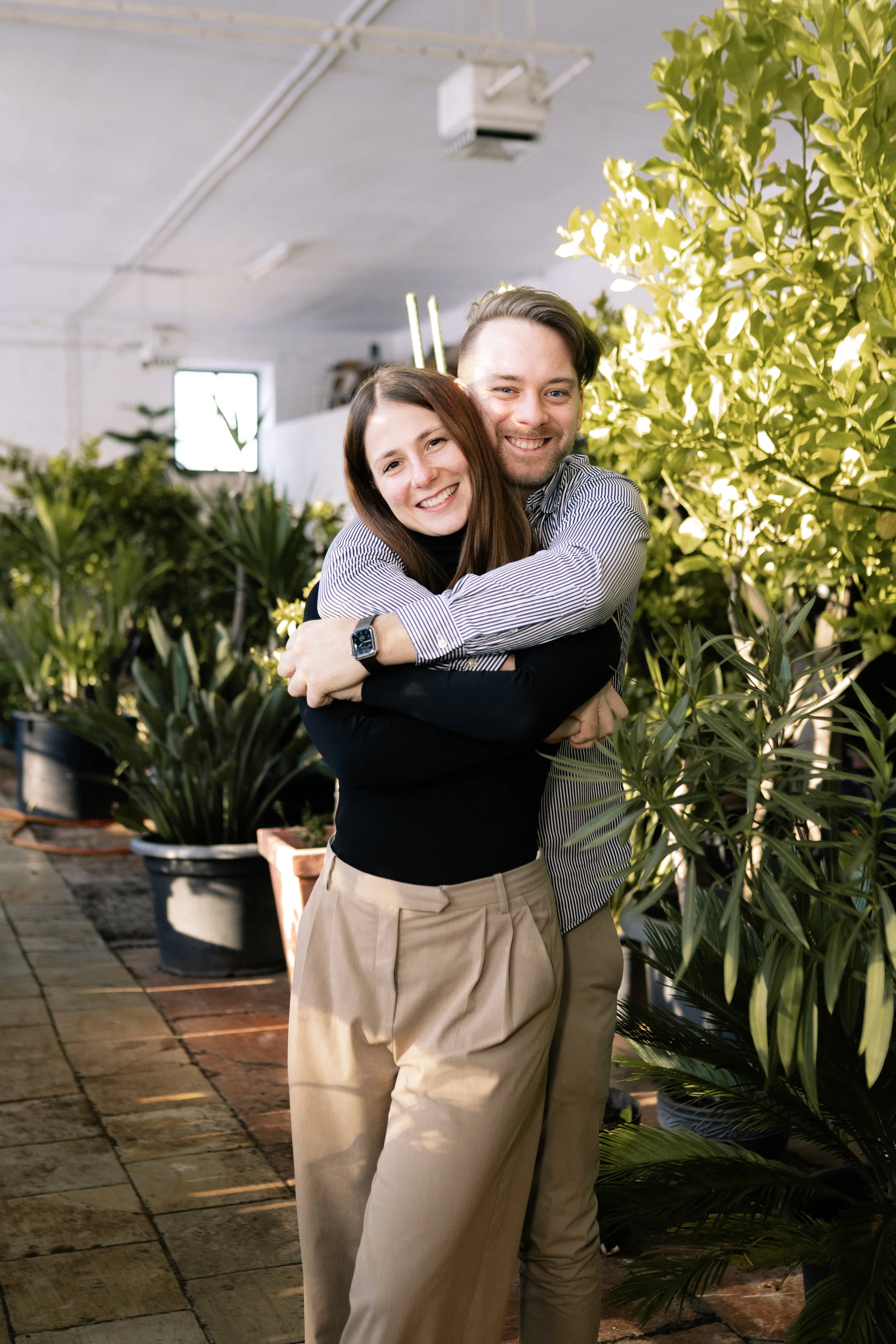 Ein lächelndes Paar umarmt sich in einem Pflanzenhaus, umgeben von verschiedenen Topfpflanzen