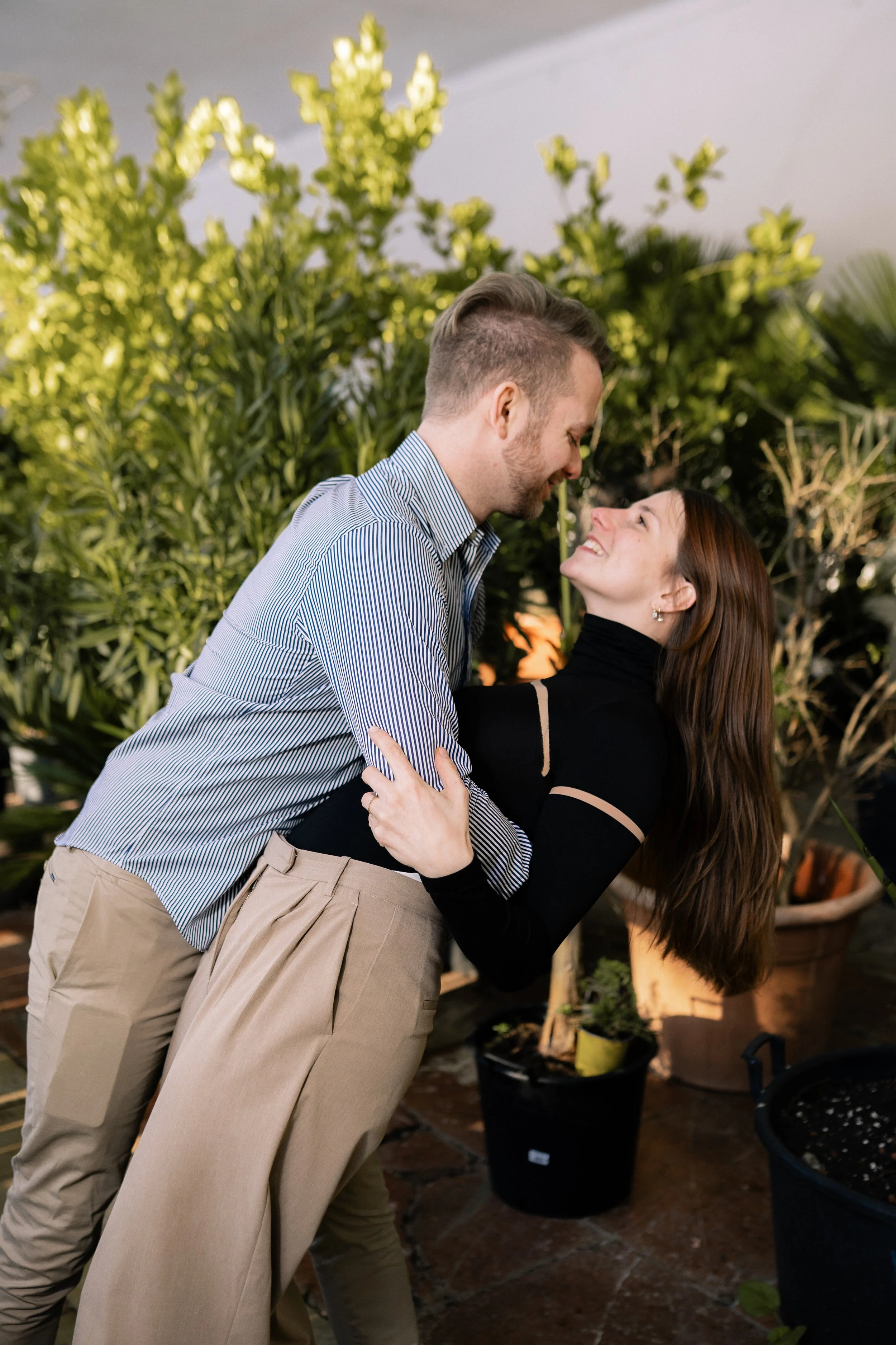 Ein lachendes Paar, das sich umarmt und sich liebevoll ansieht, auf einer Terrasse mit Pflanzen im Hintergrund.