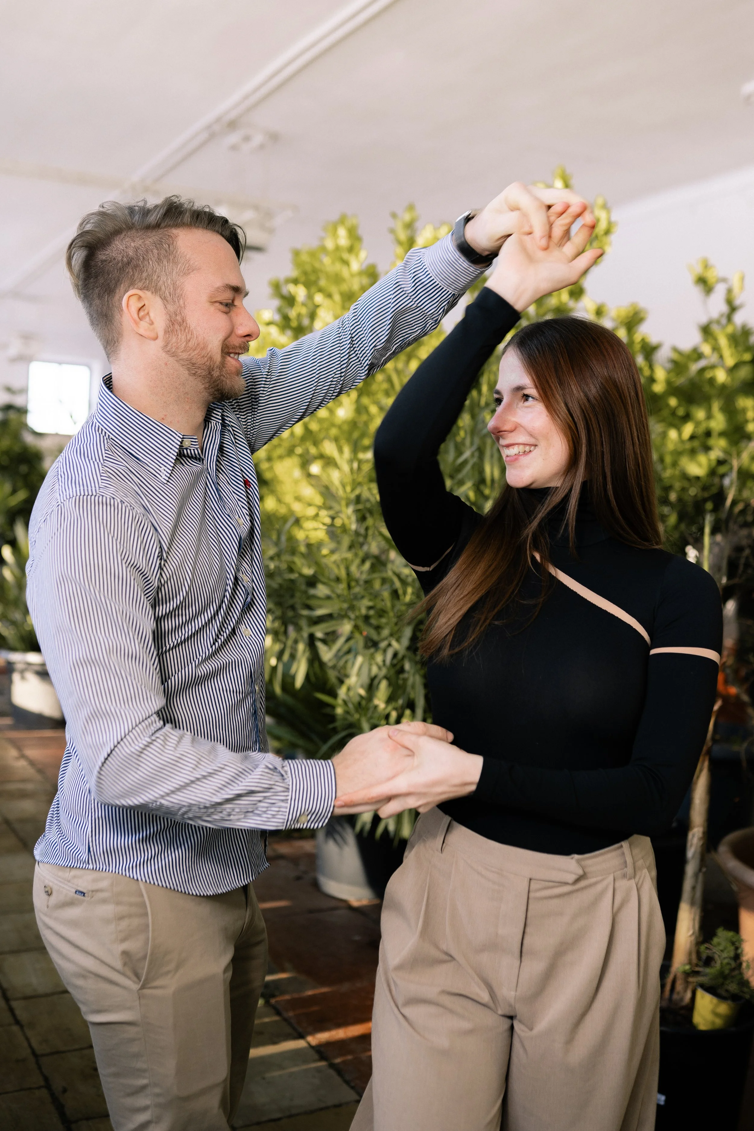 Ein Mann und eine Frau tanzen in einer Umgebung mit vielen grünen Pflanzen, sie lächeln sich an und halten sich während des Tanzes an den Händen.