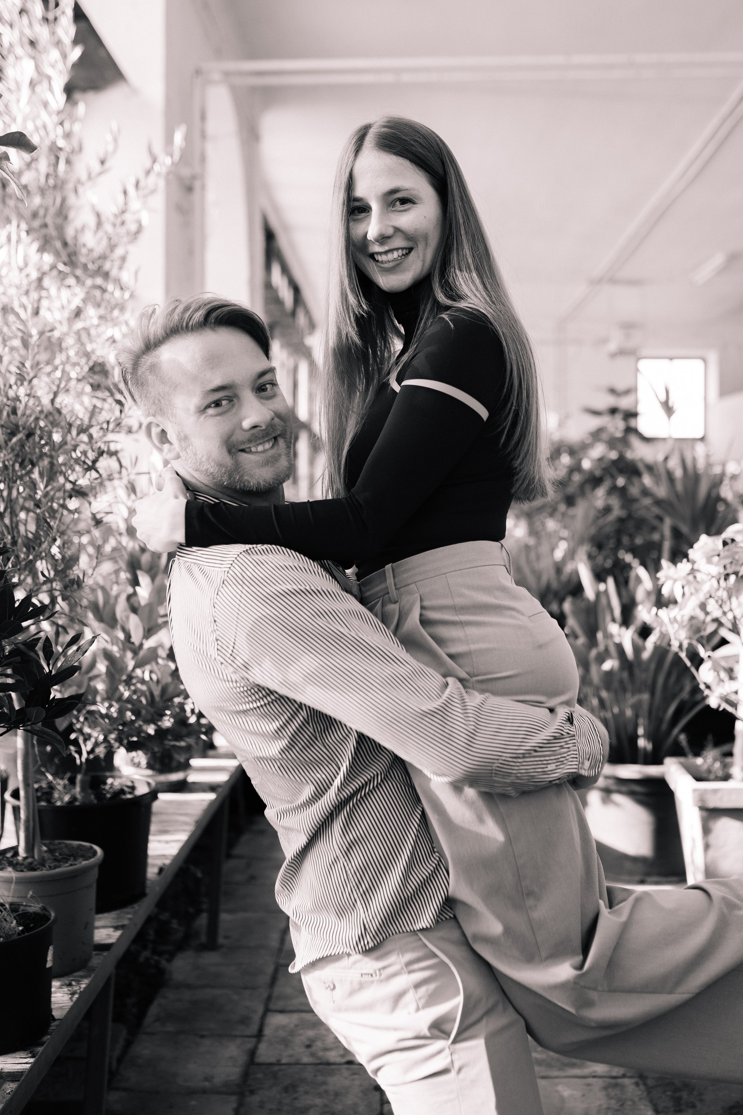 Ein Mann trägt eine Frau auf den Armen, beide lächelnd, in einem Raum mit Pflanzen im Hintergrund, schwarz-weiß