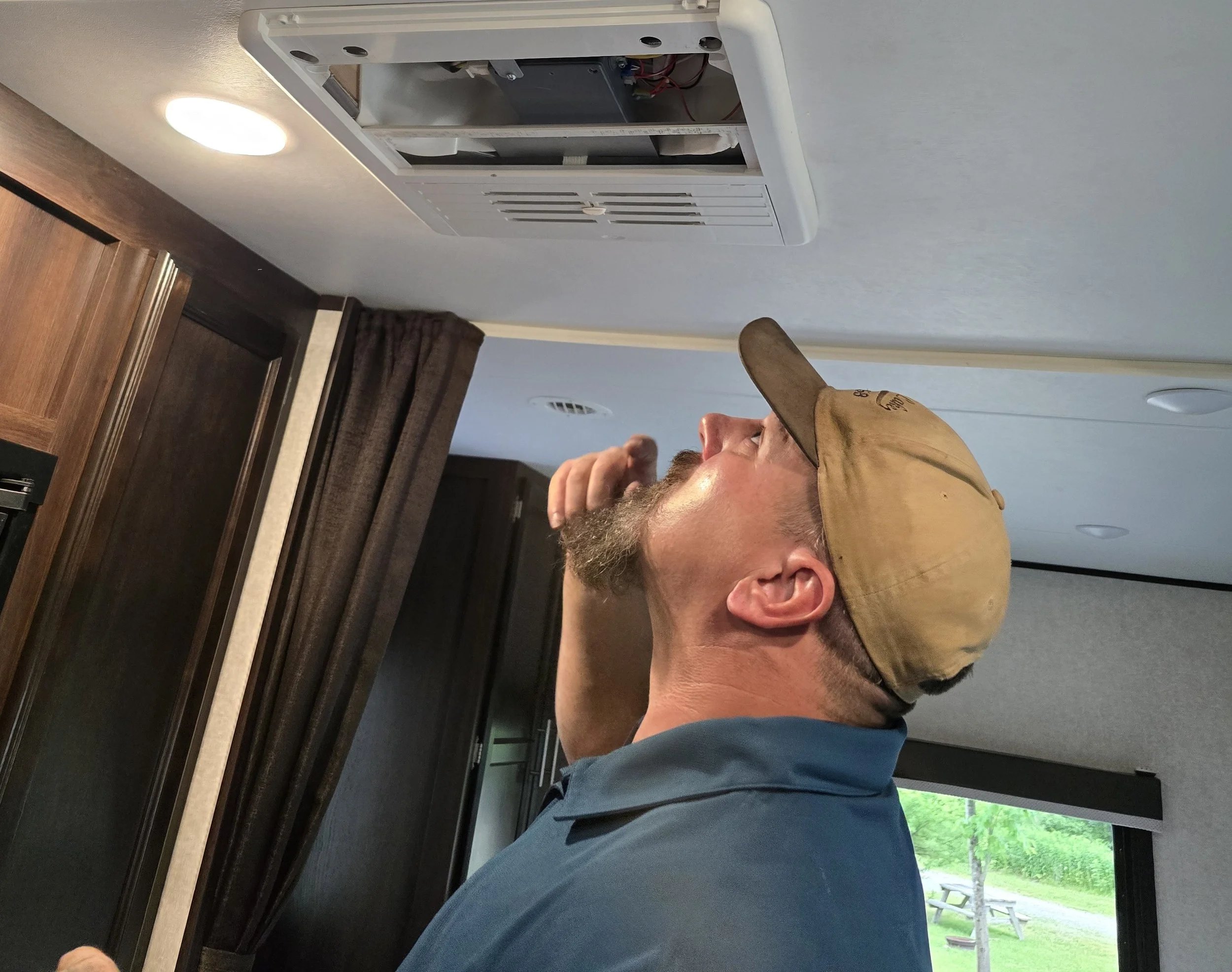 Douglas wearing a tan baseball cap and a blue shirt, standing inside a trailer or RV, looking upwards at an opening in the ceiling near an air conditioning vent.