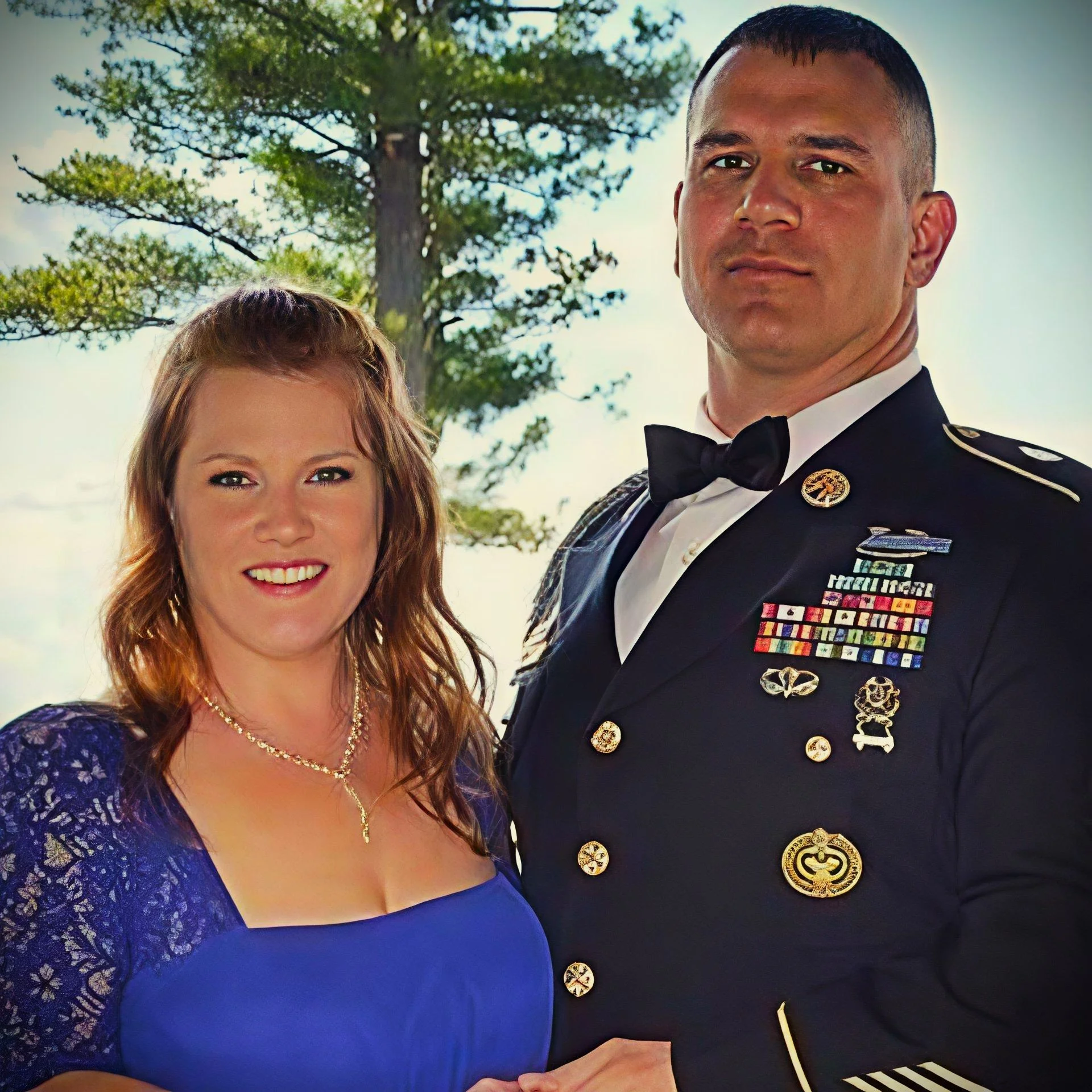 A woman in a blue dress and a man in a military uniform pose together outdoors with trees in the background.