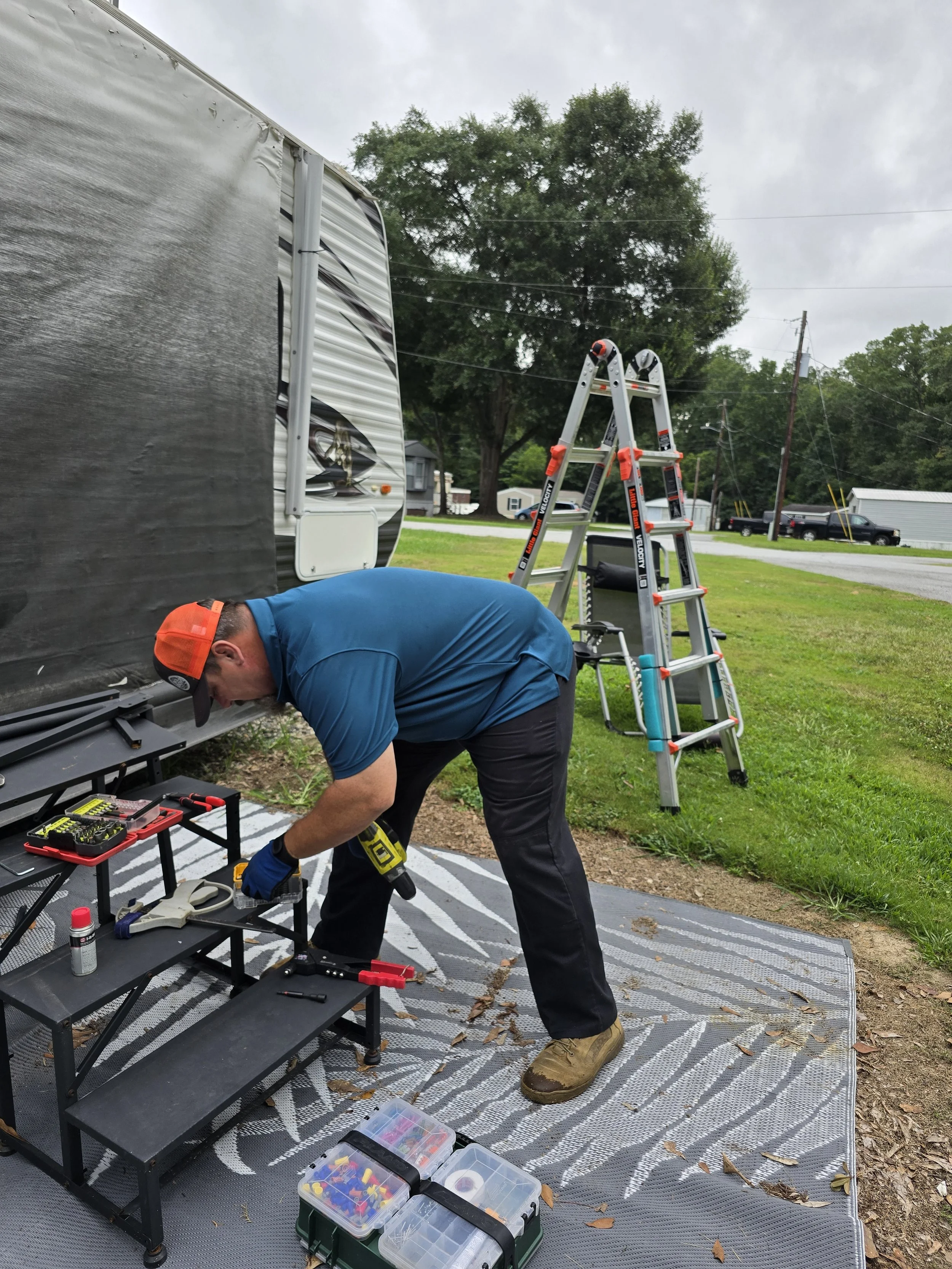 A man with a blue shirt, black pants, and brown boots working outdoors on a DIY project, using a power drill on a small black table with tools, under a black winnebago RV, with a ladder, toolbox, and trees in the background.