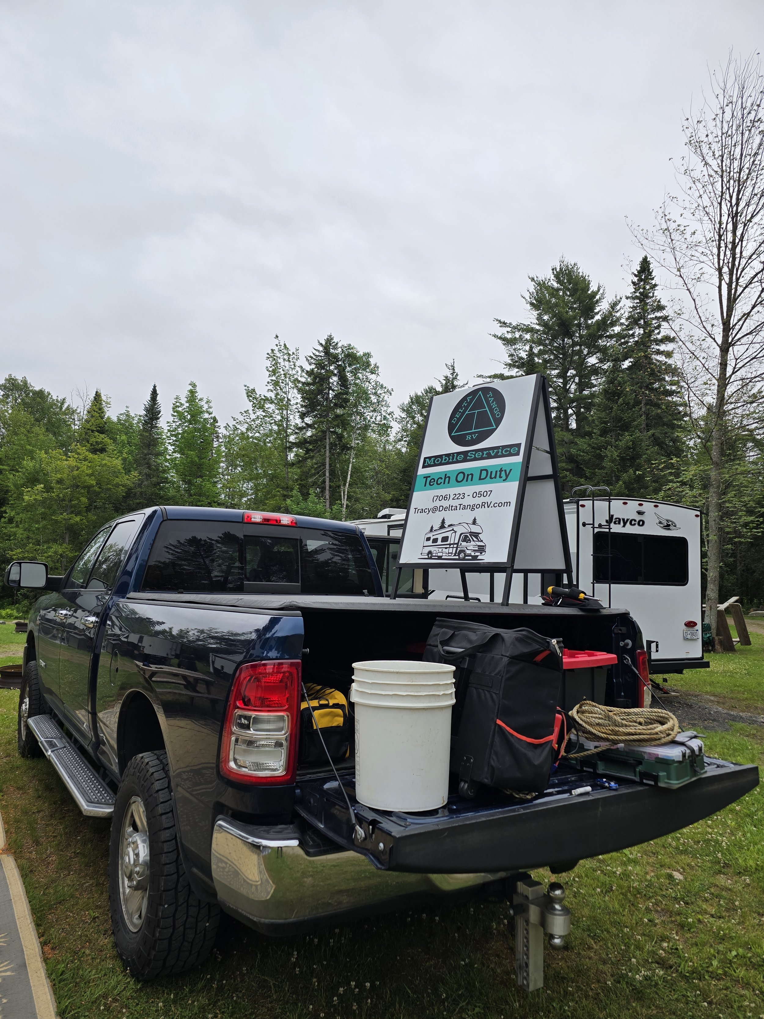 A pickup truck parked outdoors with equipment in its bed, including a white bucket, tools, and bags, in front of a travel trailer. A sign on a stand reads 'Mobile Service,' 'Tech On Duty,' with a phone number and email address, and features a graphic of a recreational vehicle.