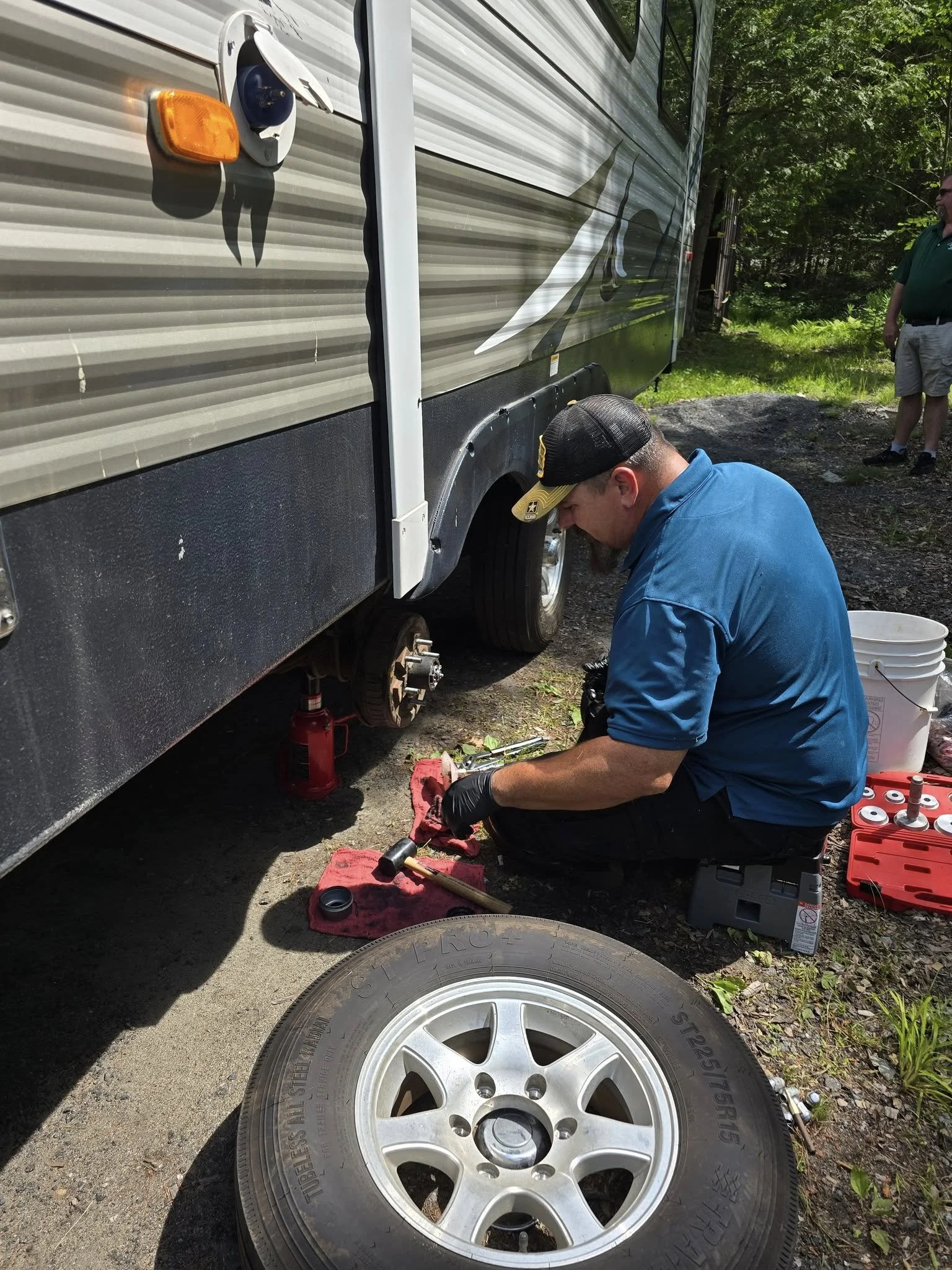 A man in a blue shirt and black gloves changing a flat tire on an RV. The RV is raised with a hydraulic jack, and the man is working near the wheel, with tools and a red toolbox nearby. Another person in a green shirt and shorts stands in the background.