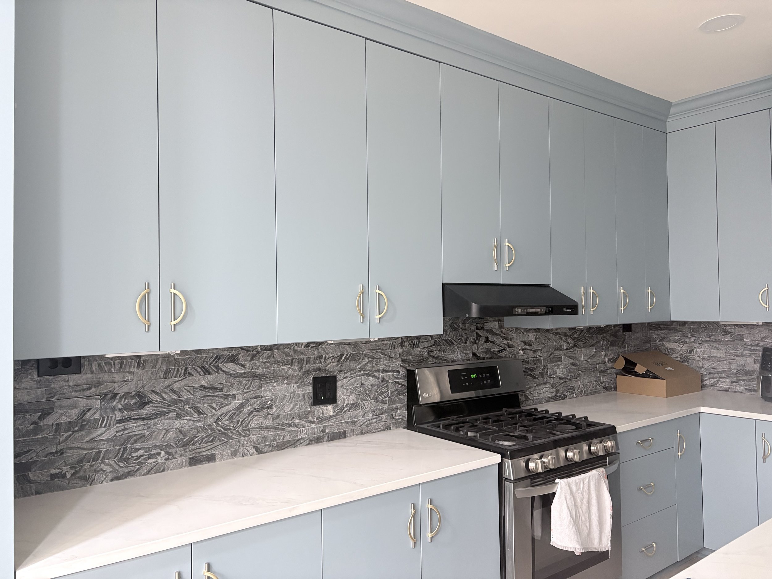 Kitchen with light blue cabinets, a black range oven, a black range hood, gray stone backsplash, white countertops, and a box on the counter.