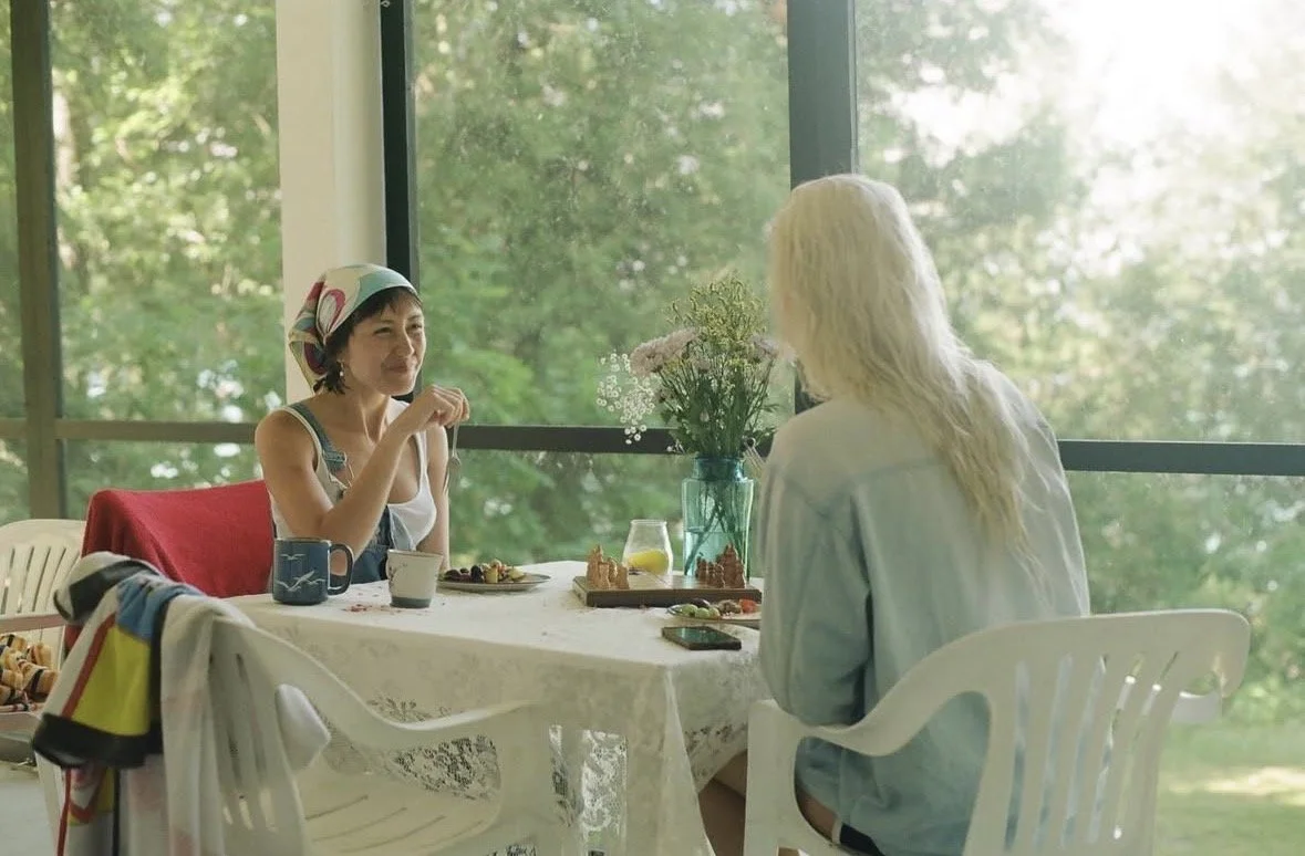 Two women having a conversation at a table in a sunlit room with large windows. One woman has short dark hair and a colorful headscarf, the other has long blonde hair. The table has flowers, drinks, and food.