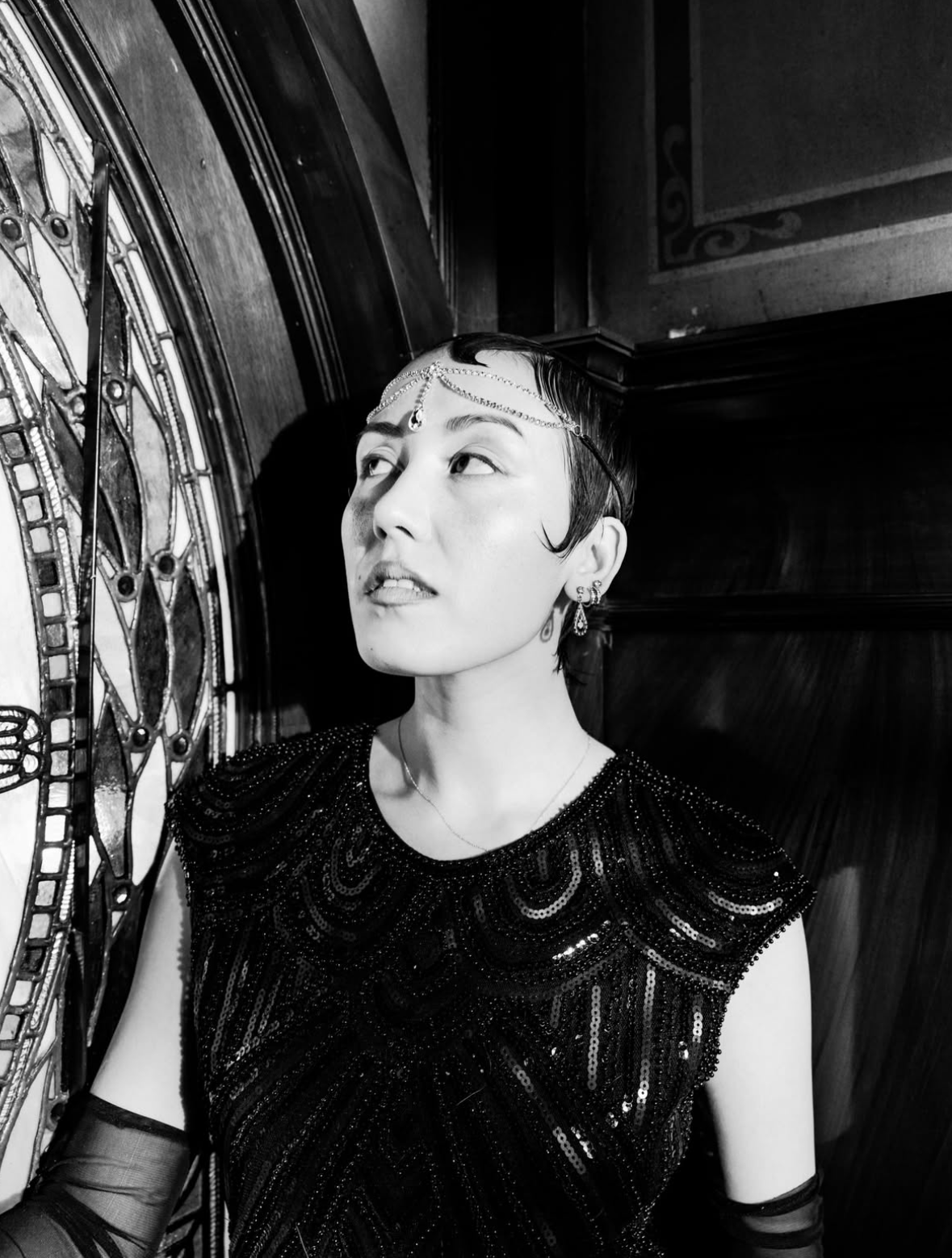 A person with short dark hair styled with waves, wearing glamorous jewelry and a black, beaded, sequined dress, posing against a stained glass window in a dimly lit, vintage-style interior.