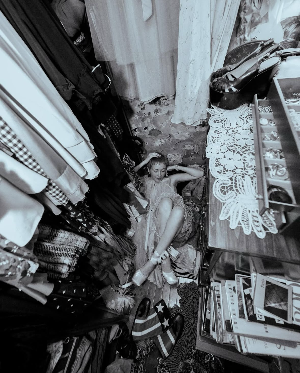 A young woman lies on the floor in a cluttered room, surrounded by clothing, accessories, and books, with her arms resting on her head and her eyes closed.