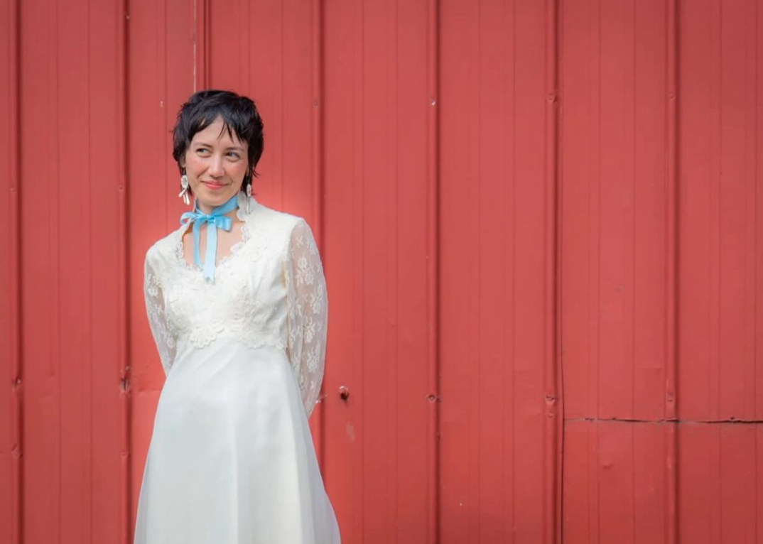 Woman with short dark hair wearing a white lace dress and a blue scarf around her neck, standing against a red wooden wall.