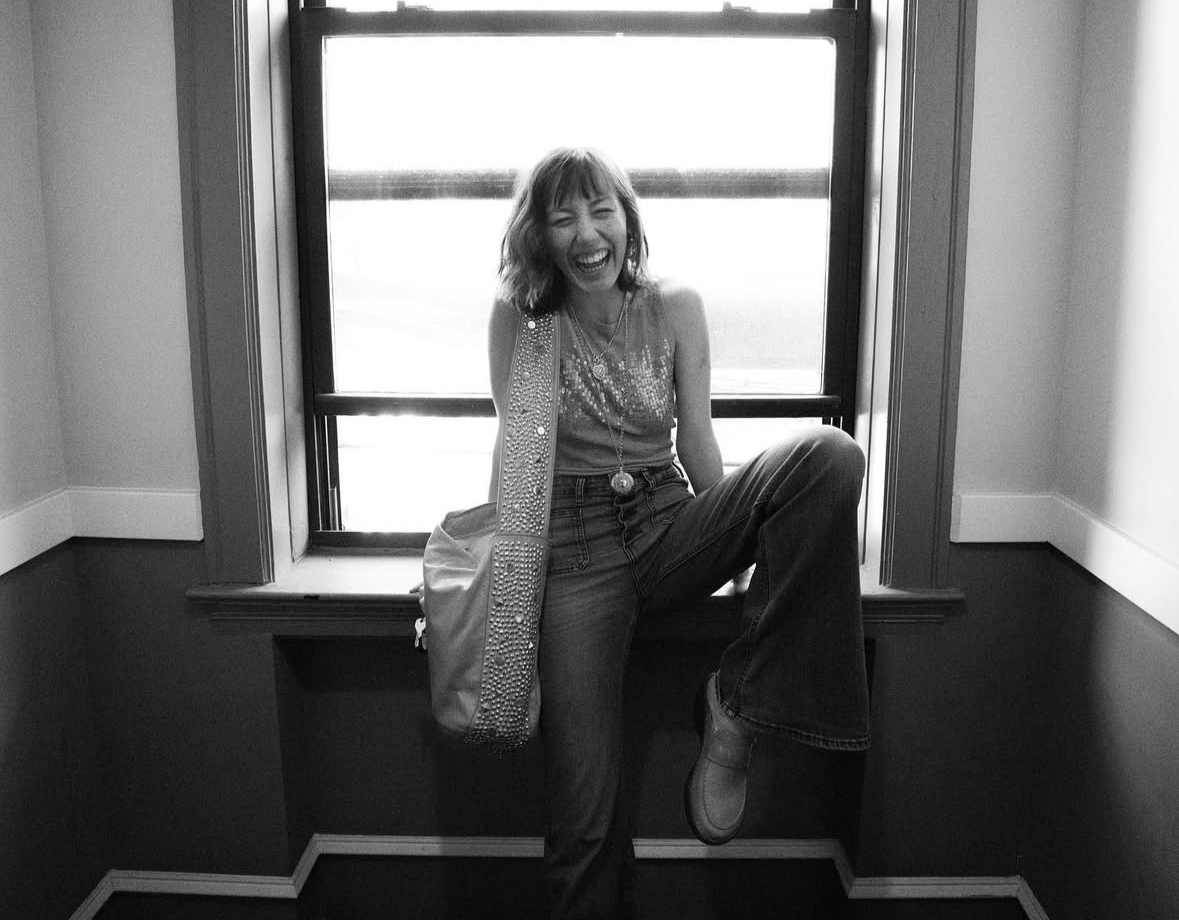 A woman sitting on a windowsill, laughing and smiling, in a room with light-colored walls and dark wainscoting.