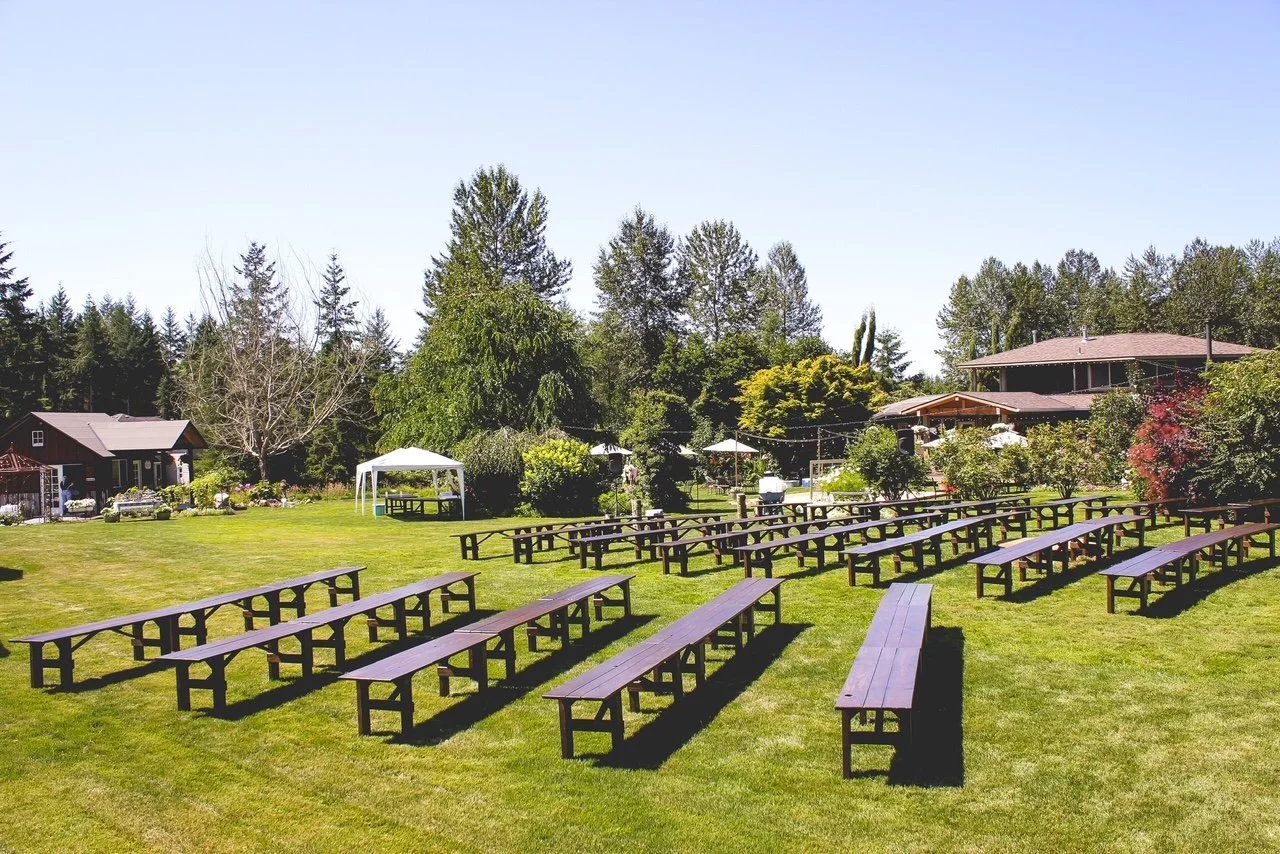 Outdoor scene with many wooden picnic tables arranged on a well-maintained grassy lawn, surrounded by trees and houses, under a clear blue sky.