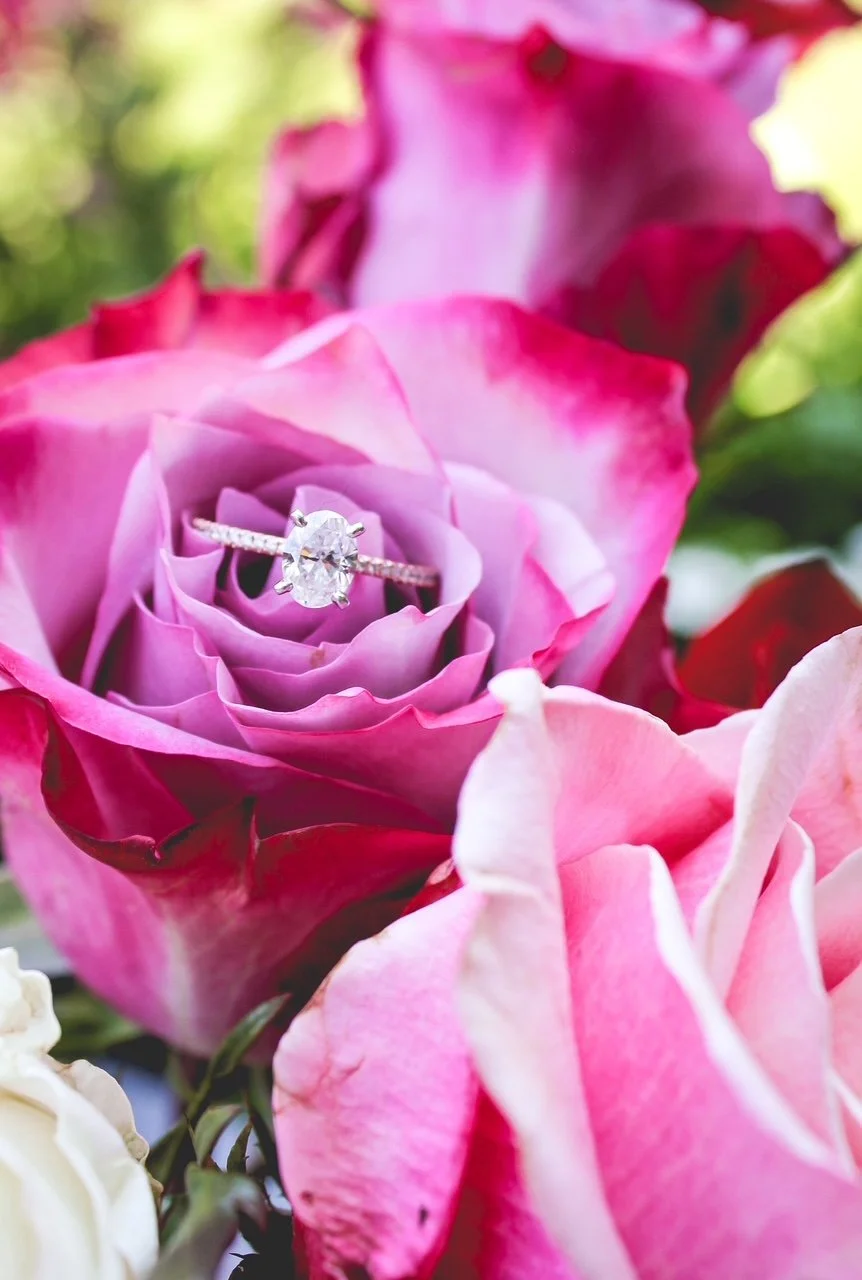 An engagement ring with a large oval diamond set in a band adorned with smaller diamonds, placed inside the petals of pink and purple roses.