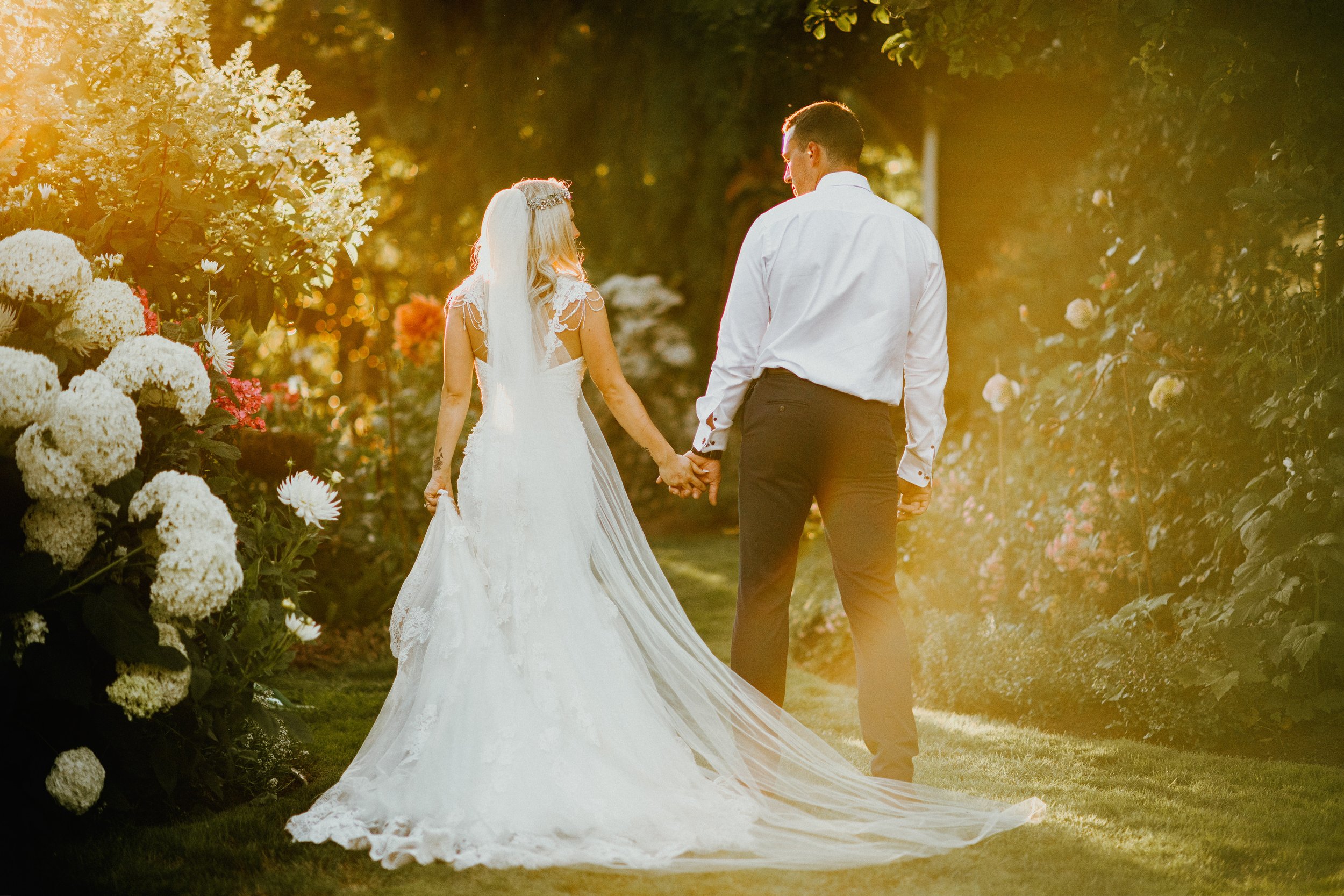 A bride and groom holding hands, walking through a garden at sunset, with flowers and greenery surrounding them.