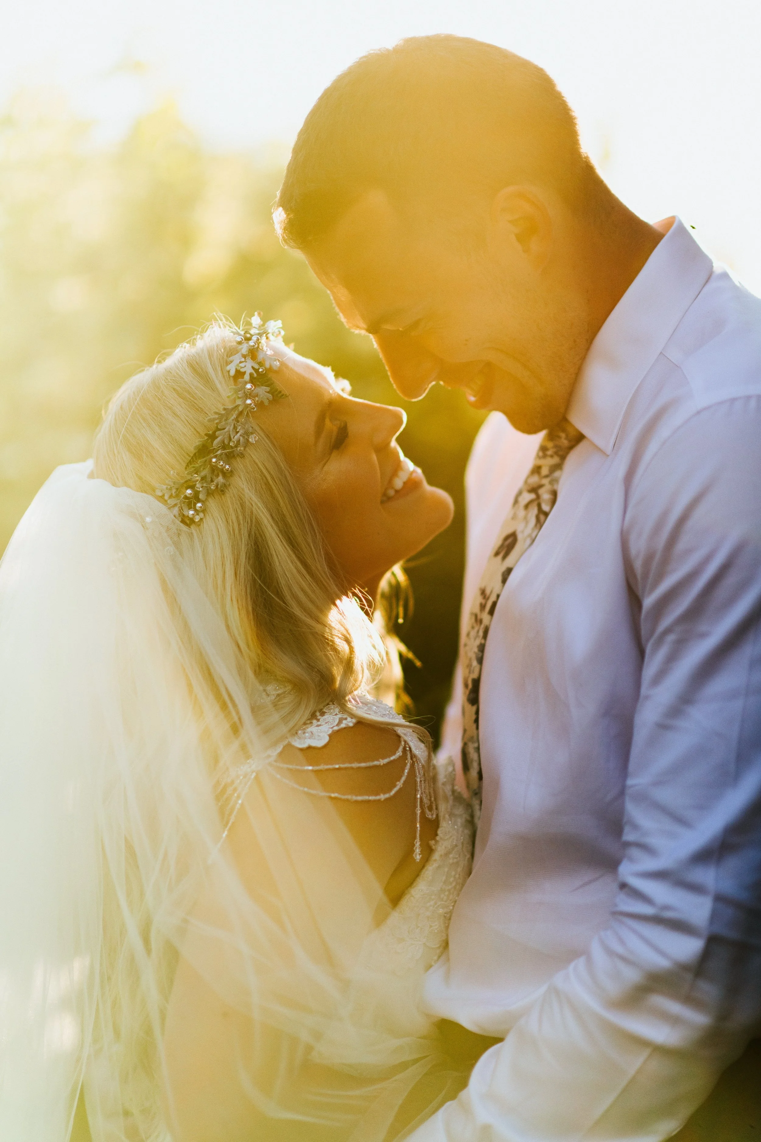 A bride and groom smiling and looking at each other outdoors during sunset, with the bride wearing a floral headband and veil, and the groom in a white shirt and patterned tie.
