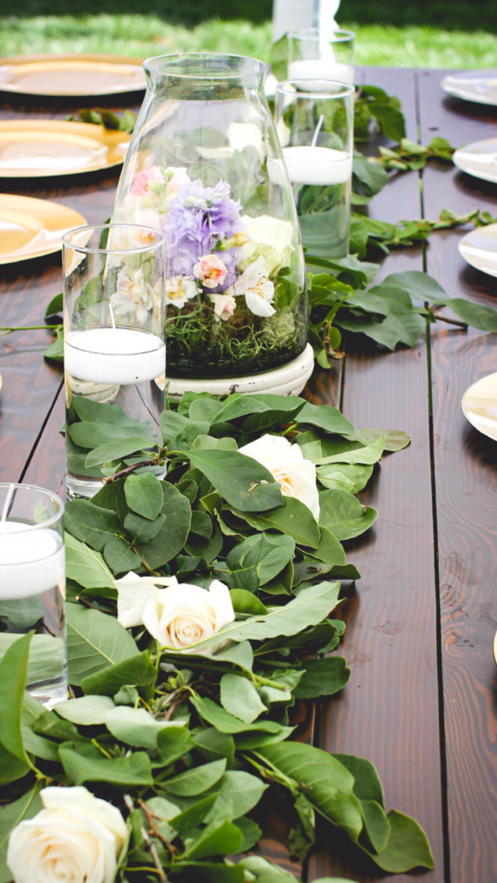 A wooden dining table decorated with a garland of green leaves and white roses, with glass candle holders and a large glass terrarium filled with lavender and other flowers as a centerpiece.