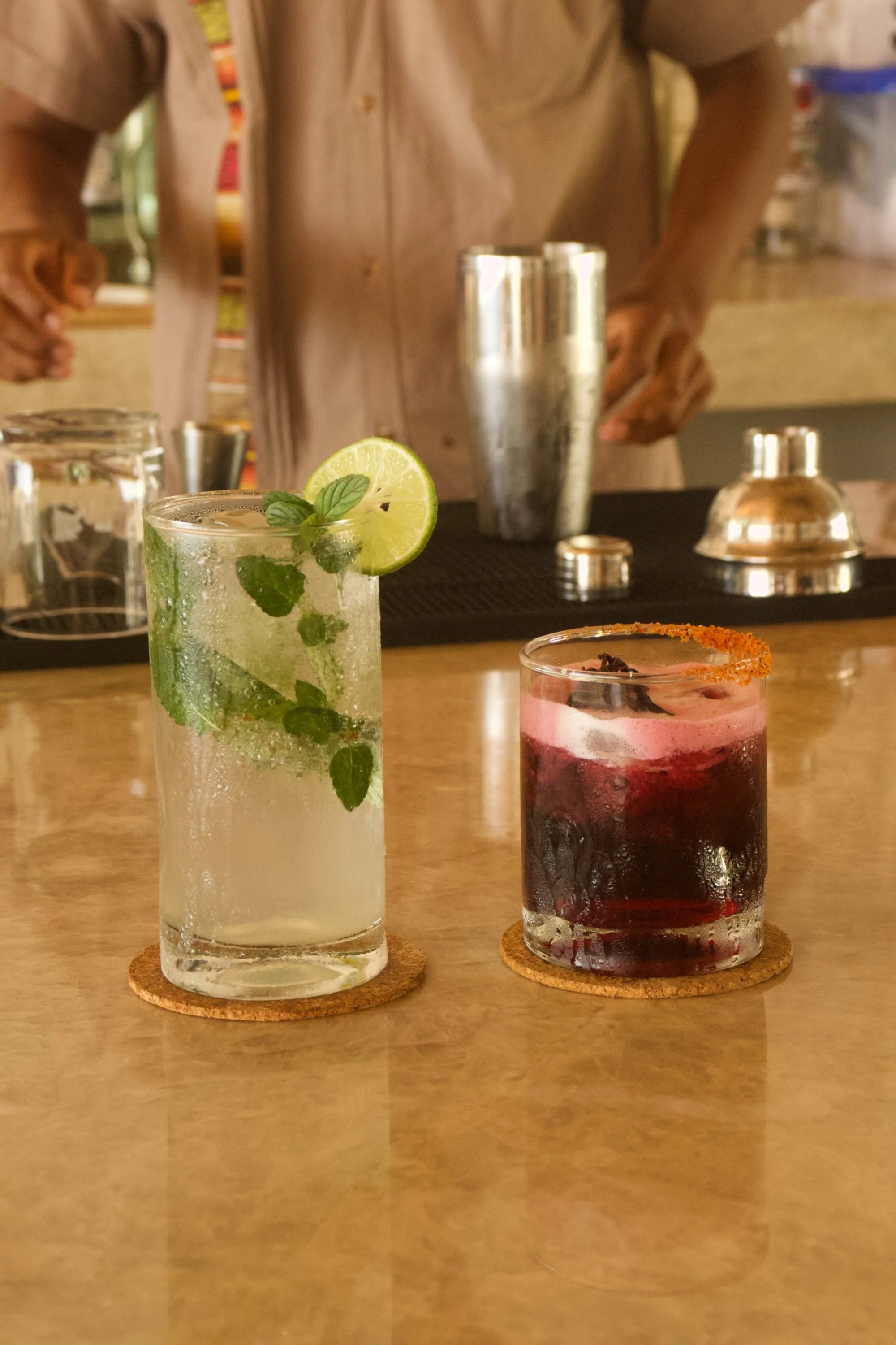 Two cocktails on a bar counter, one with lime and mint, the other with a red garnish and an orange rim, with bartenders in the background.