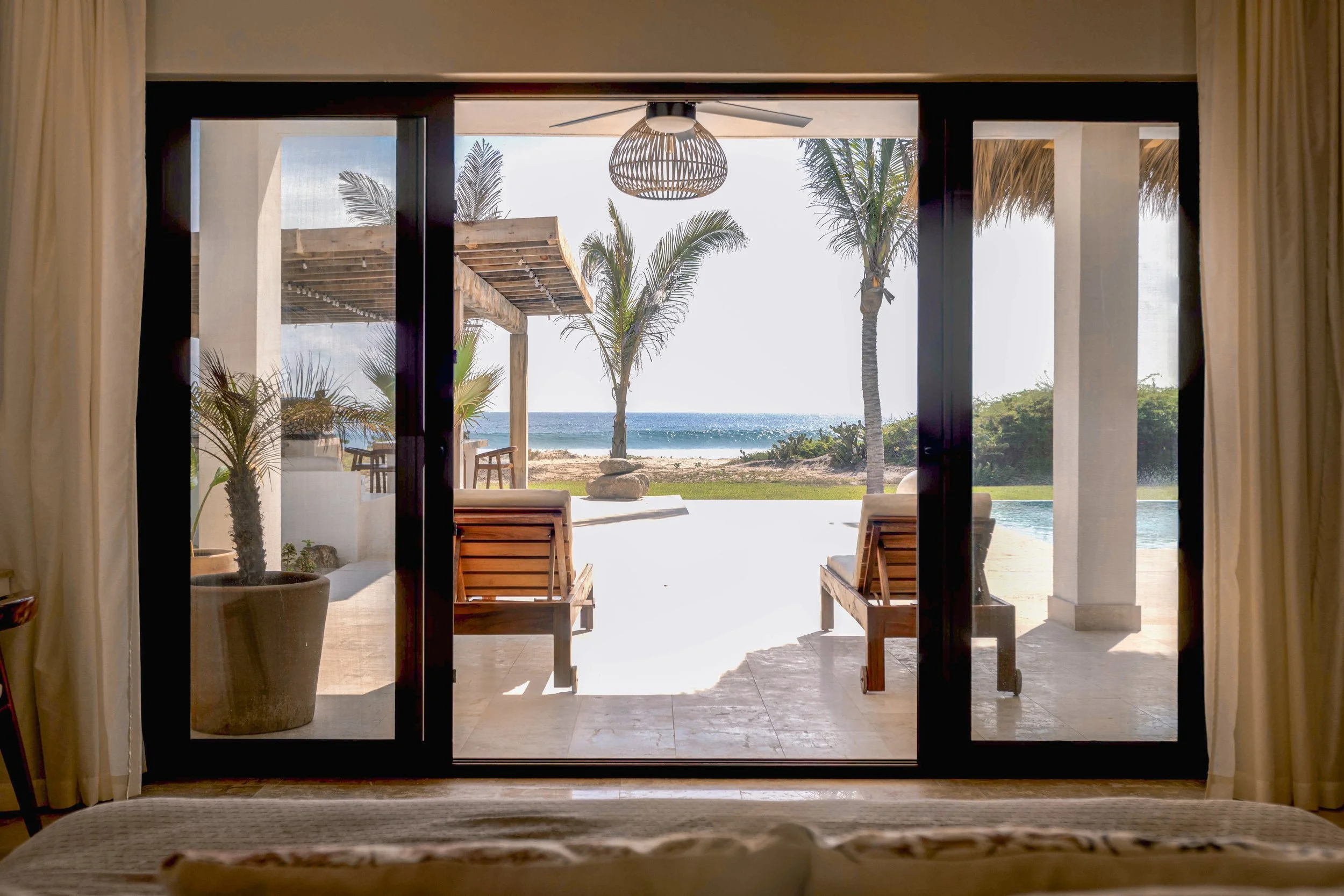 View of a tropical beach seen through open sliding glass doors from inside a room. Outdoor patio with wooden lounge chairs, palm trees, sandy beach, and ocean in the background.