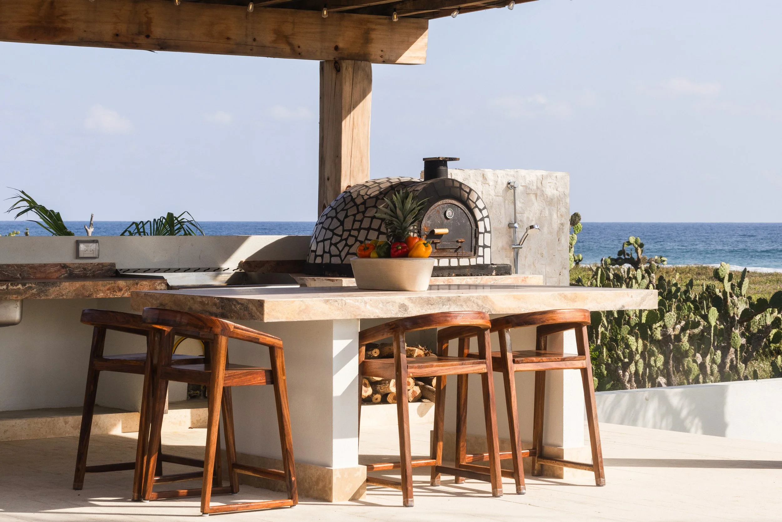 Outdoor kitchen with a wood-fired pizza oven, a bowl of fresh fruit including pineapple and bell peppers, a countertop with wooden stools, and a view of the ocean and cacti in the background.