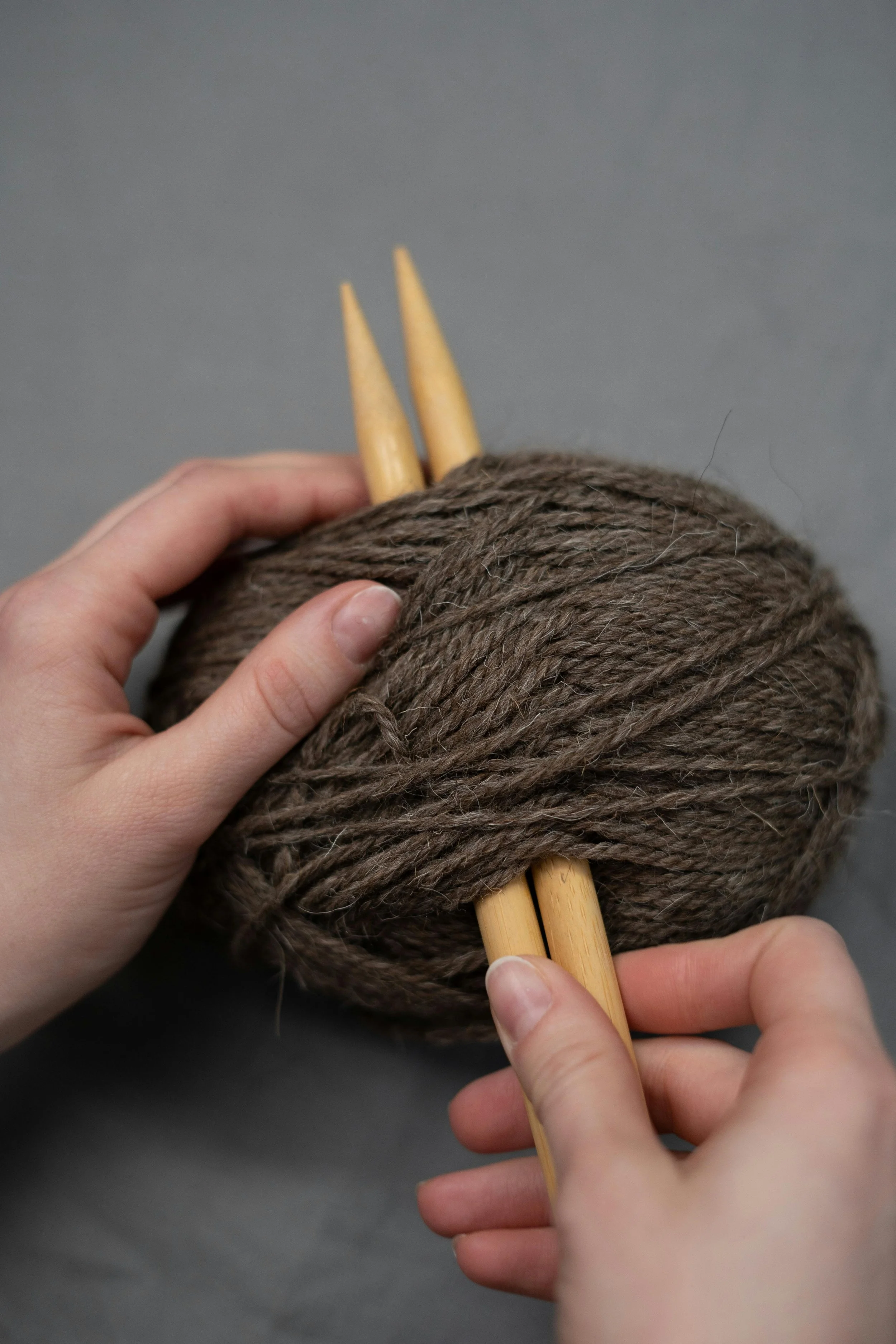 A ball of brown yarn with two knitting needles inserted, being held by two hands.