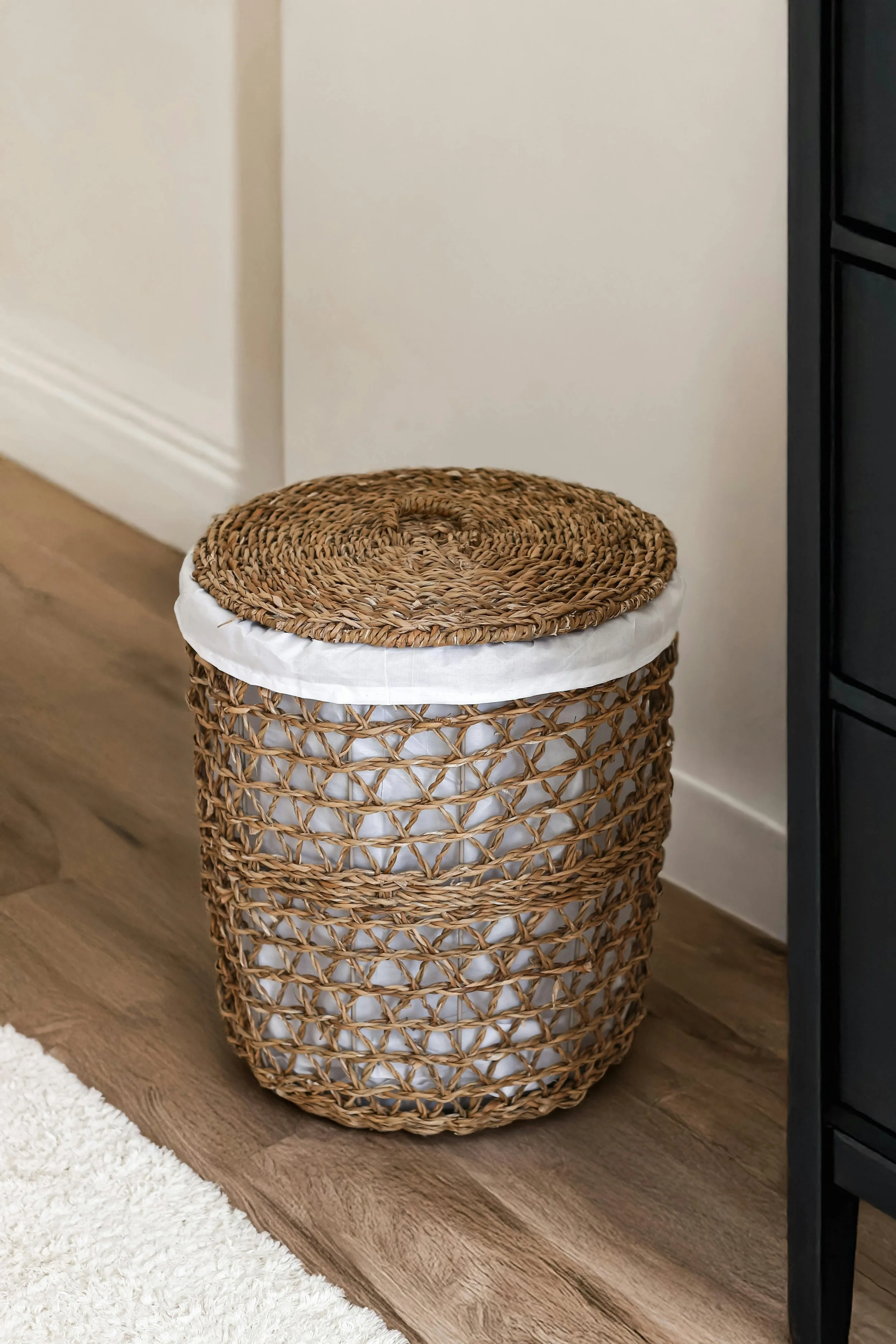 Wicker laundry basket with a cloth lining sitting on a wooden floor next to a black piece of furniture.