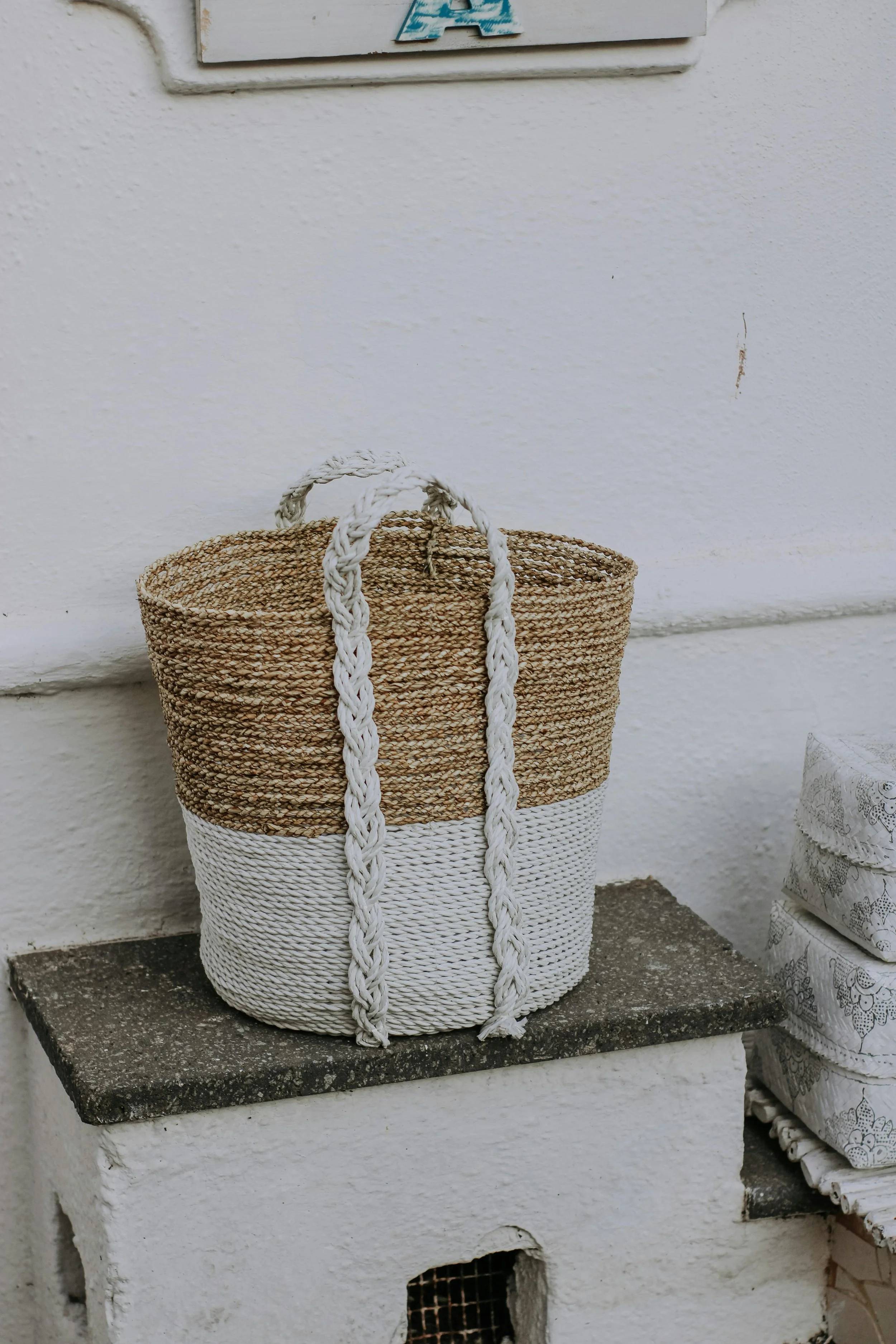A woven basket with a two-tone design, located on a white textured surface against a white wall. The basket has braided white handles and is partly sitting on a small white structure.