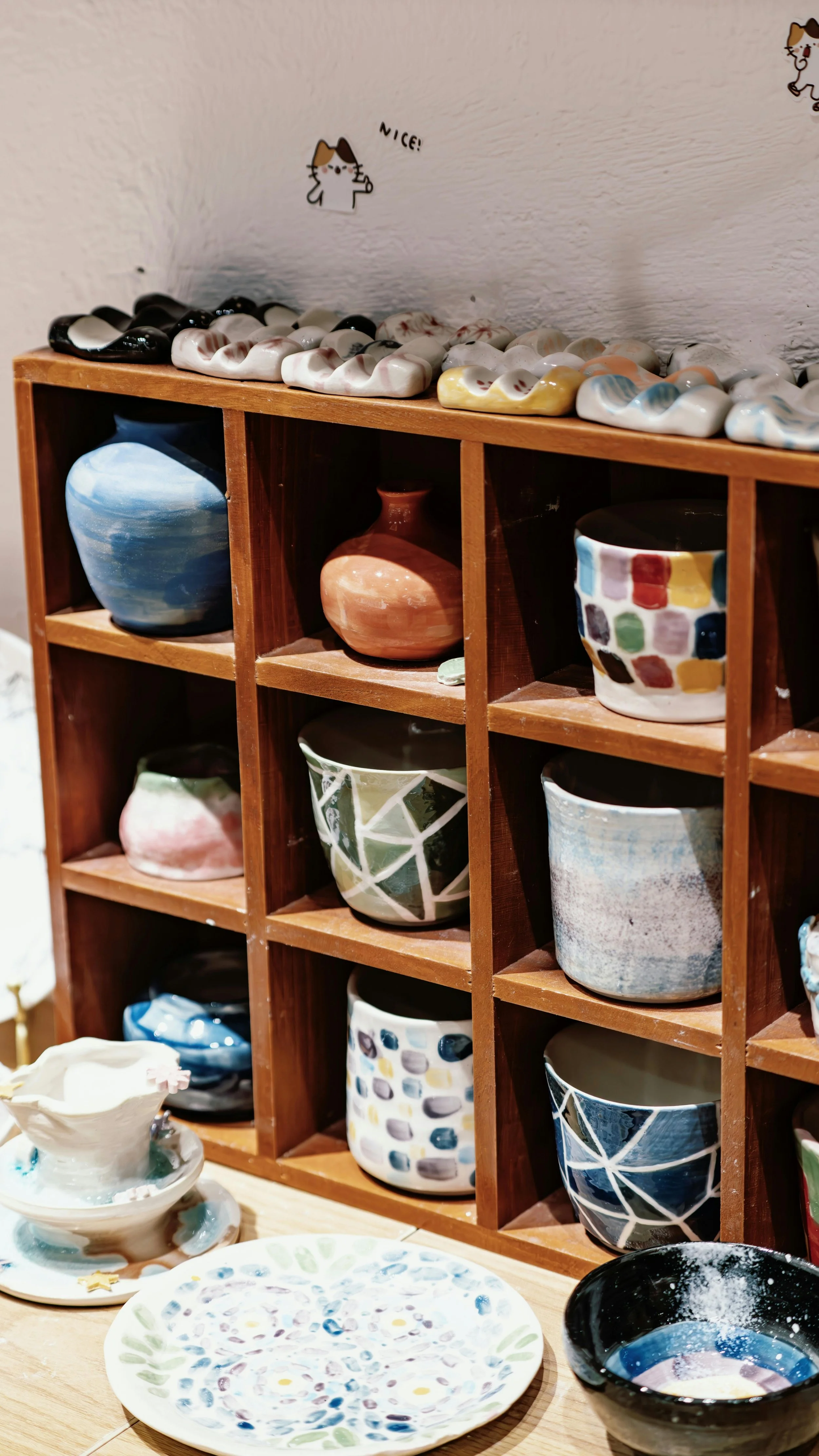 Display of colorful handmade pottery bowls and plates on a wooden shelf, with additional bowls and plates on the table in front.
