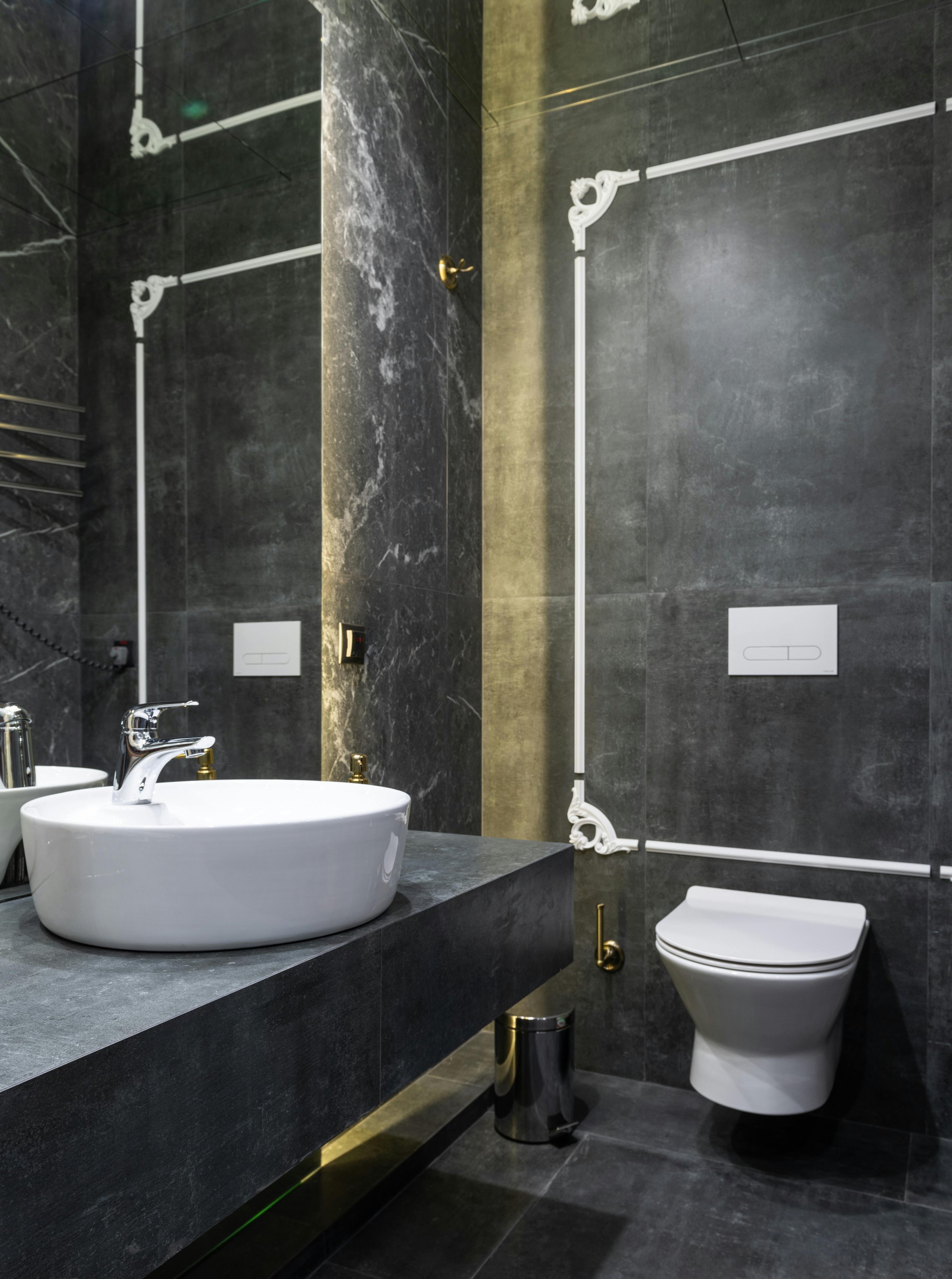 A modern bathroom with dark marble walls and flooring, featuring a white countertop with a vessel sink, gold fixtures, and a wall-mounted toilet. Decorative white piping runs along the wall, and there is a suitcase on the floor next to the toilet.