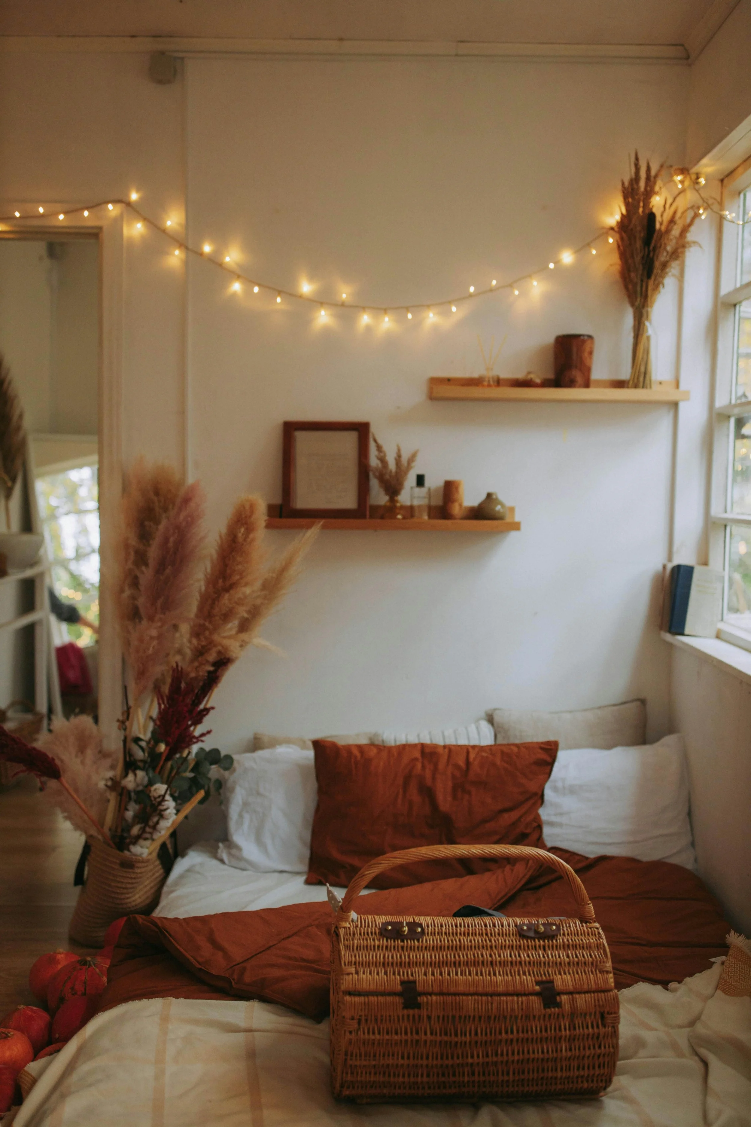 Cozy bedroom with string lights, wooden shelves decorated with dried flowers and vases, a window allowing natural light, a basket with plants, and a bed with white and rust-colored bedding.