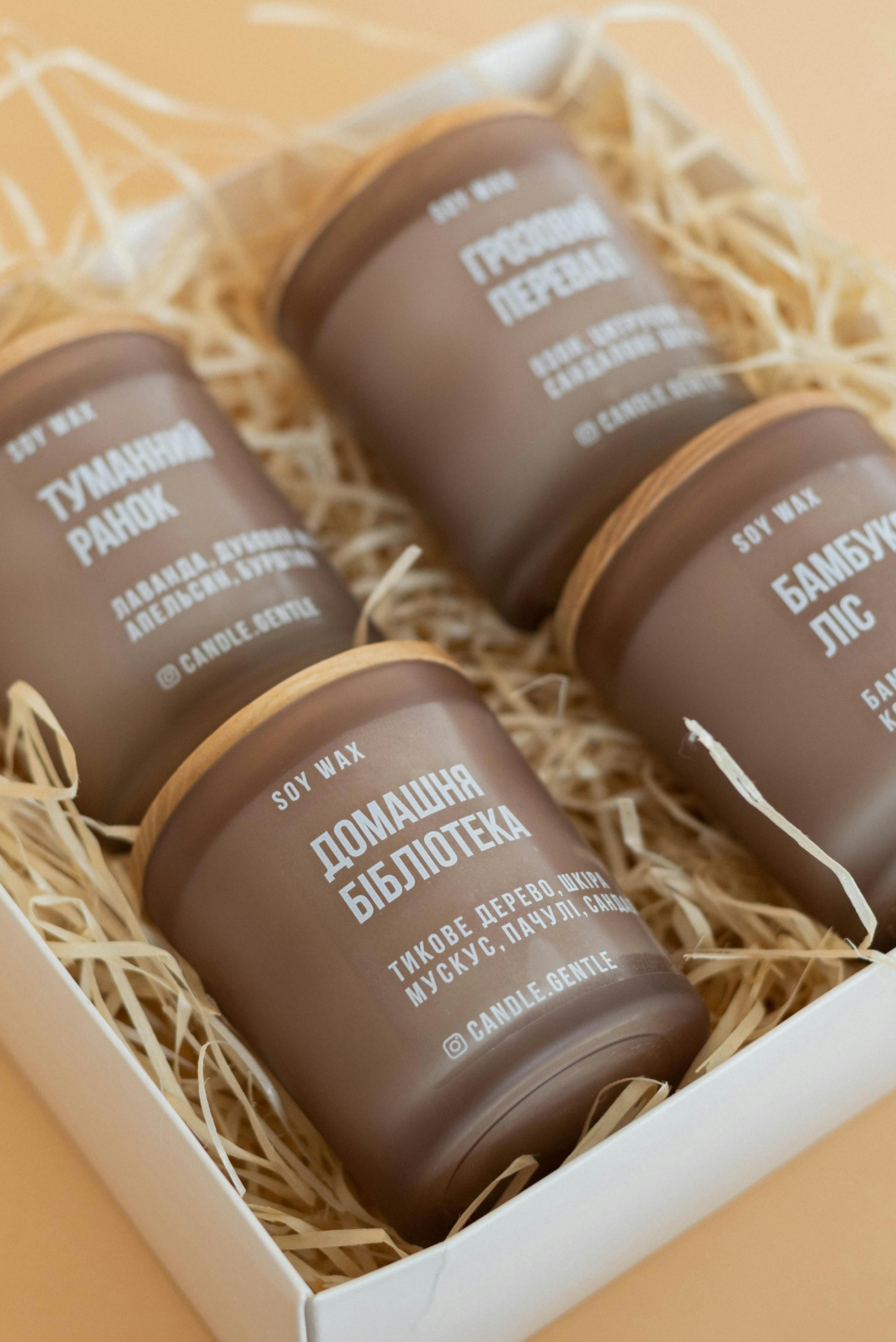 Four brown soy wax candles in glass containers with wooden lids, labeled in multiple languages, placed in a white box with shredded paper filler.