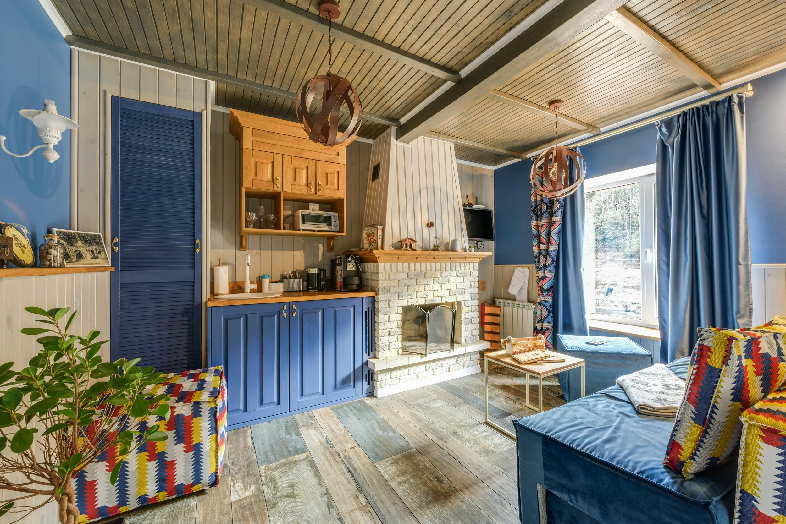 Living room with a blue cabinet next to a brick fireplace, a colorful patterned armchair, a small coffee table, blue curtains, and a large window showing a snowy outdoor scene.