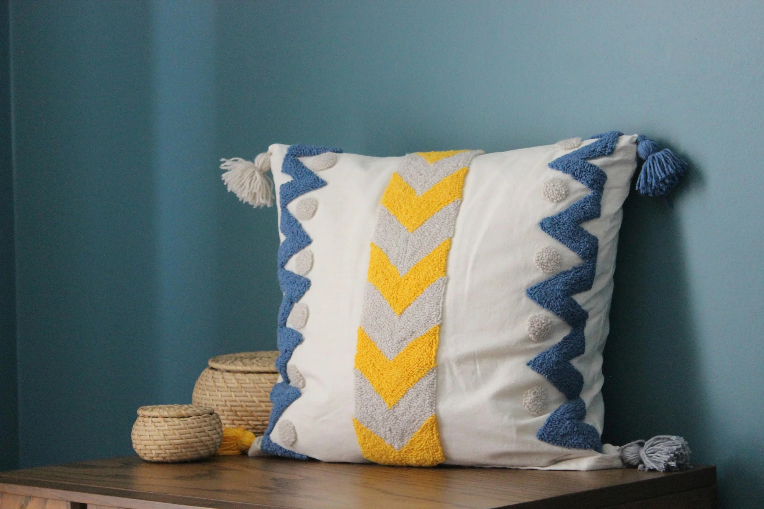 Decorative white pillow with blue, gray, and yellow chevron and pom-pom trim on a wooden surface against a blue wall, accompanied by two woven baskets.