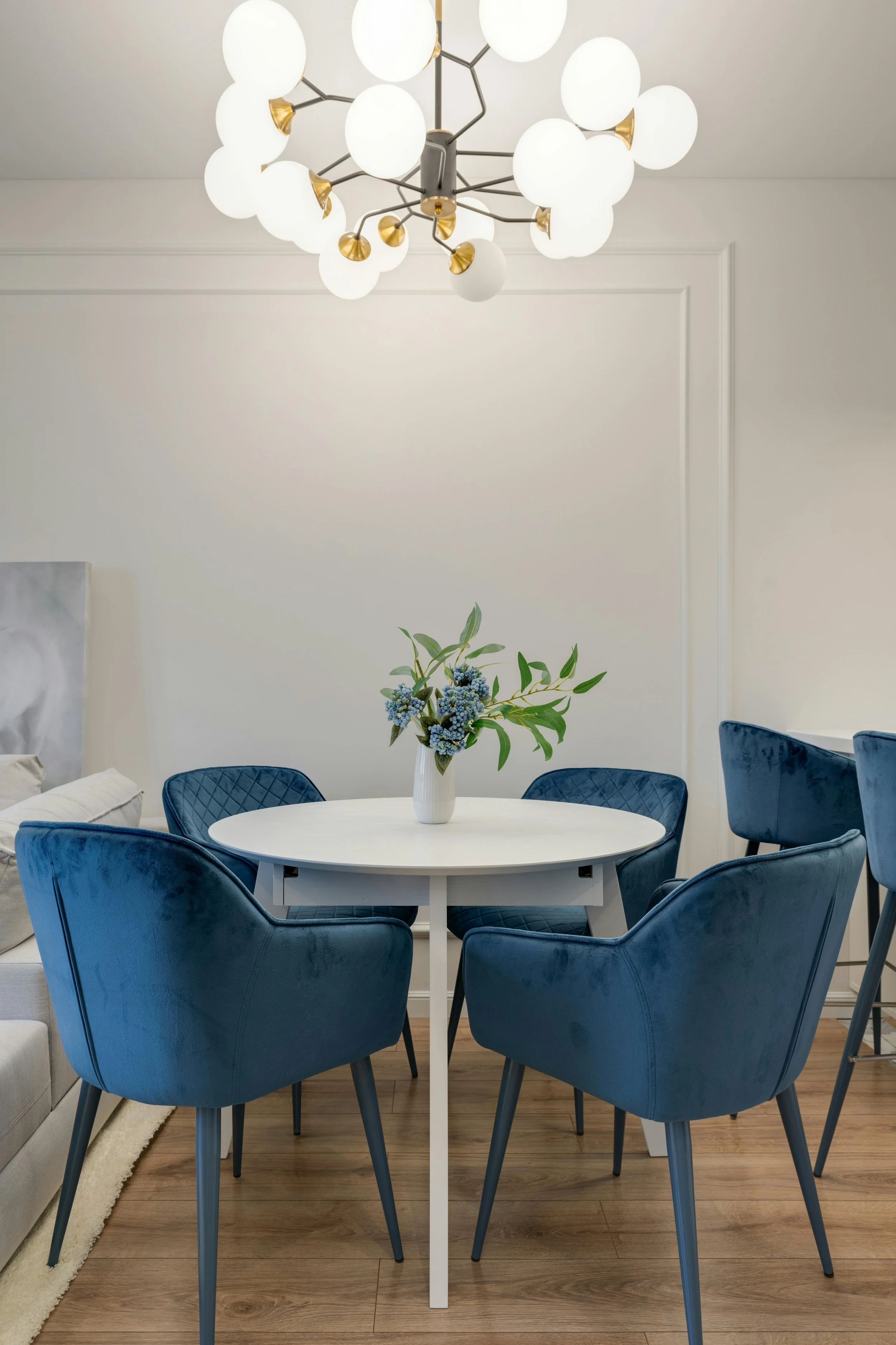 White round dining table with six dark blue velvet upholstered chairs arranged around it, a white vase with blue and green flowers, and a modern chandelier with white globes and gold accents hanging from the ceiling in a bright room with white walls and wooden floor.